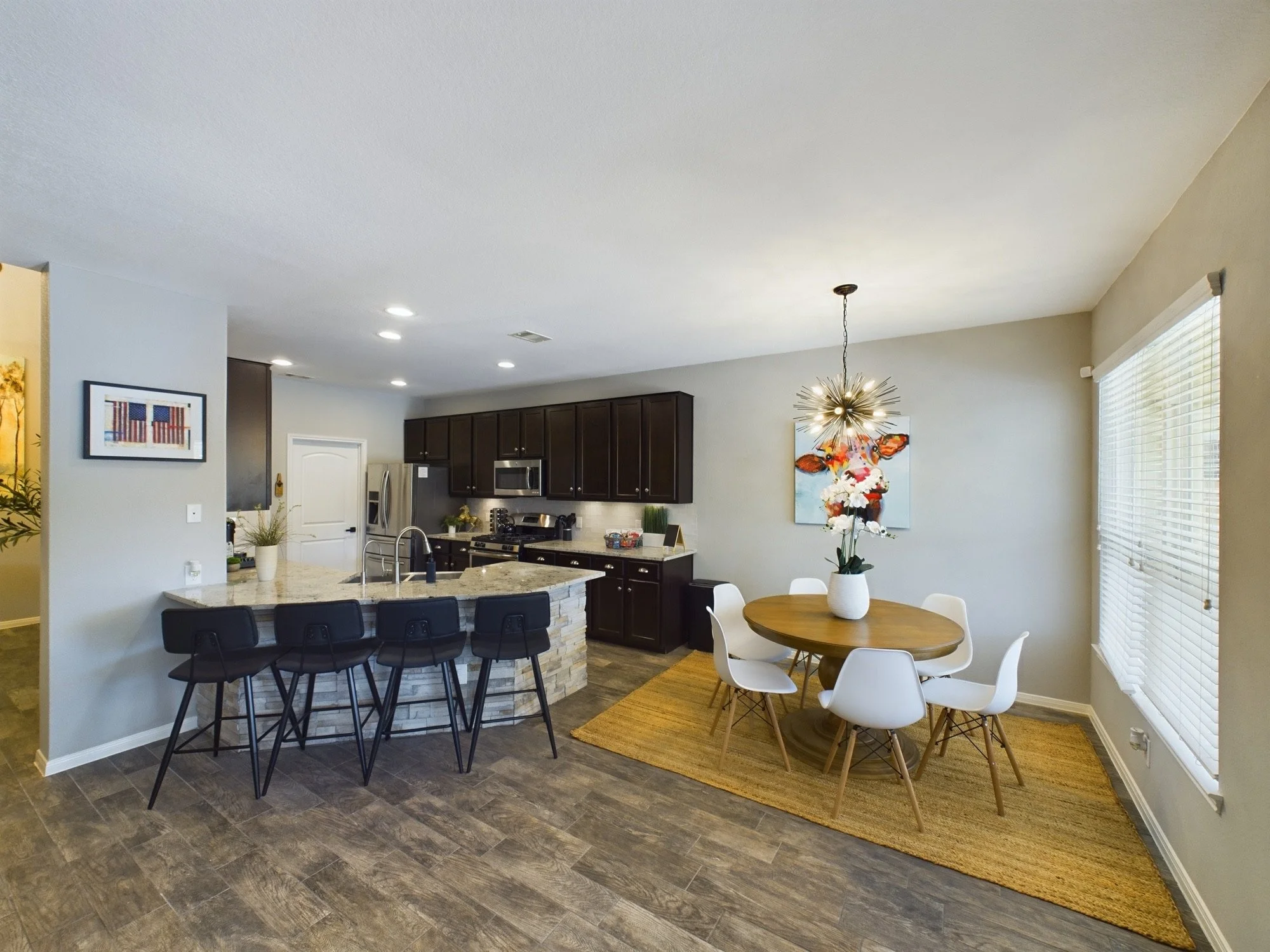 Open-concept kitchen and dining area with dark wood cabinets, stainless steel appliances, a marble countertop island, and a round wooden dining table with white chairs, beige walls, a floral painting, and a modern chandelier.