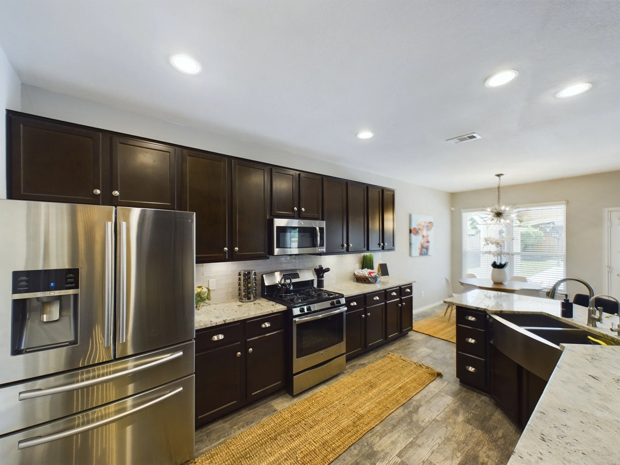 Modern kitchen with stainless steel refrigerator, dark wood cabinets, granite countertops, and a dining area with a window and chandelier.