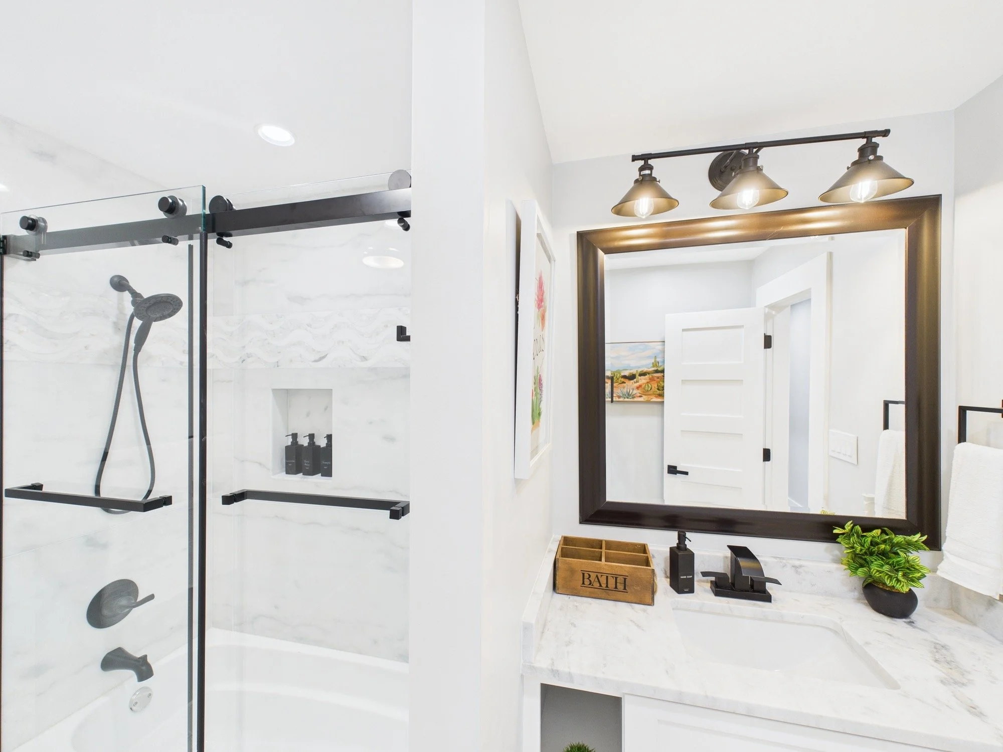 Modern bathroom with a walk-in shower, white marble walls, black accents, and a large mirror with three industrial-style light fixtures above it.