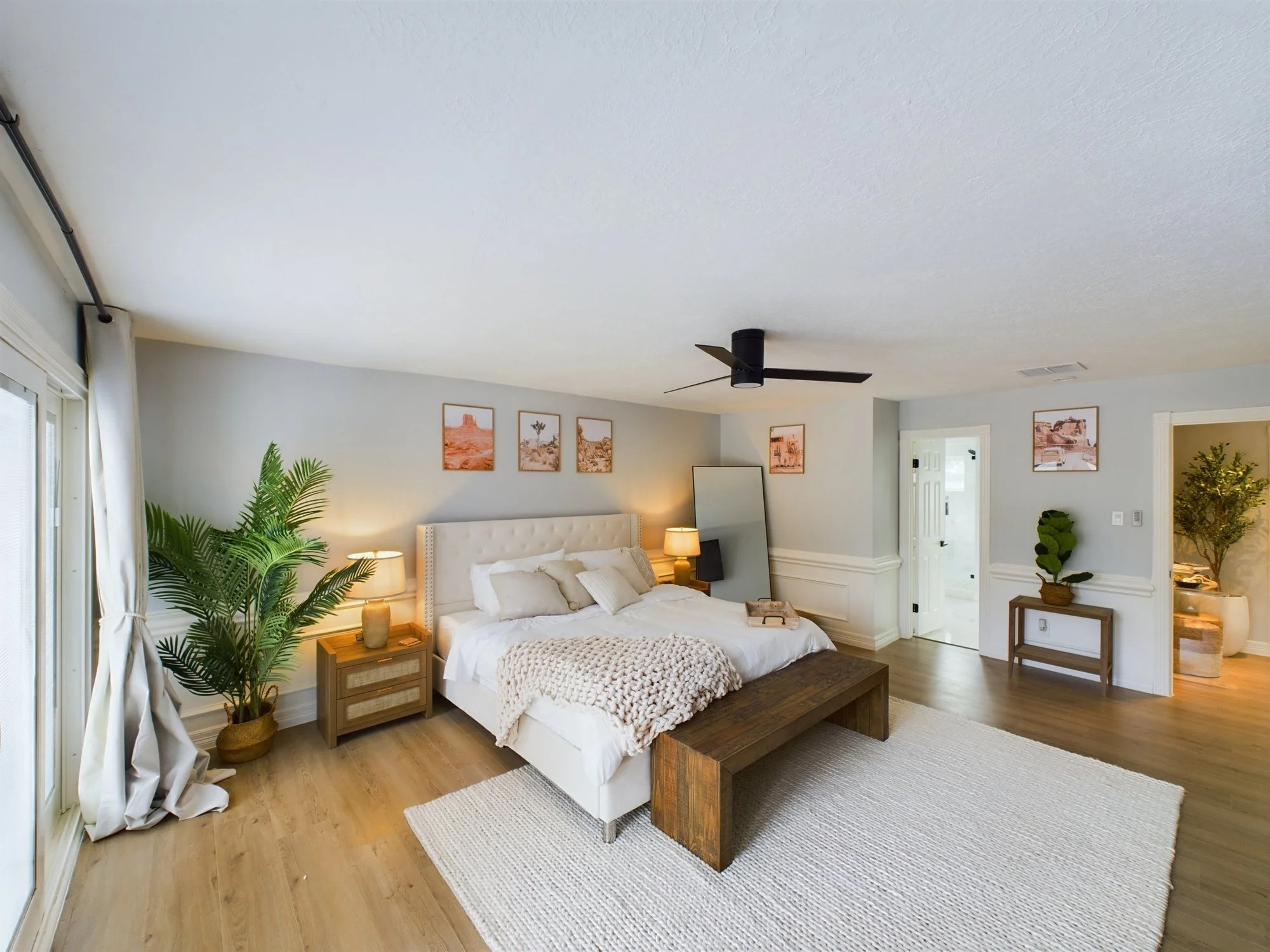 A modern bedroom with white walls, a white bed with beige and gray pillows, and a chunky beige blanket. There are nightstands with lamps on each side of the bed. A large potted plant is near the window, and three framed desert landscape photos hang a