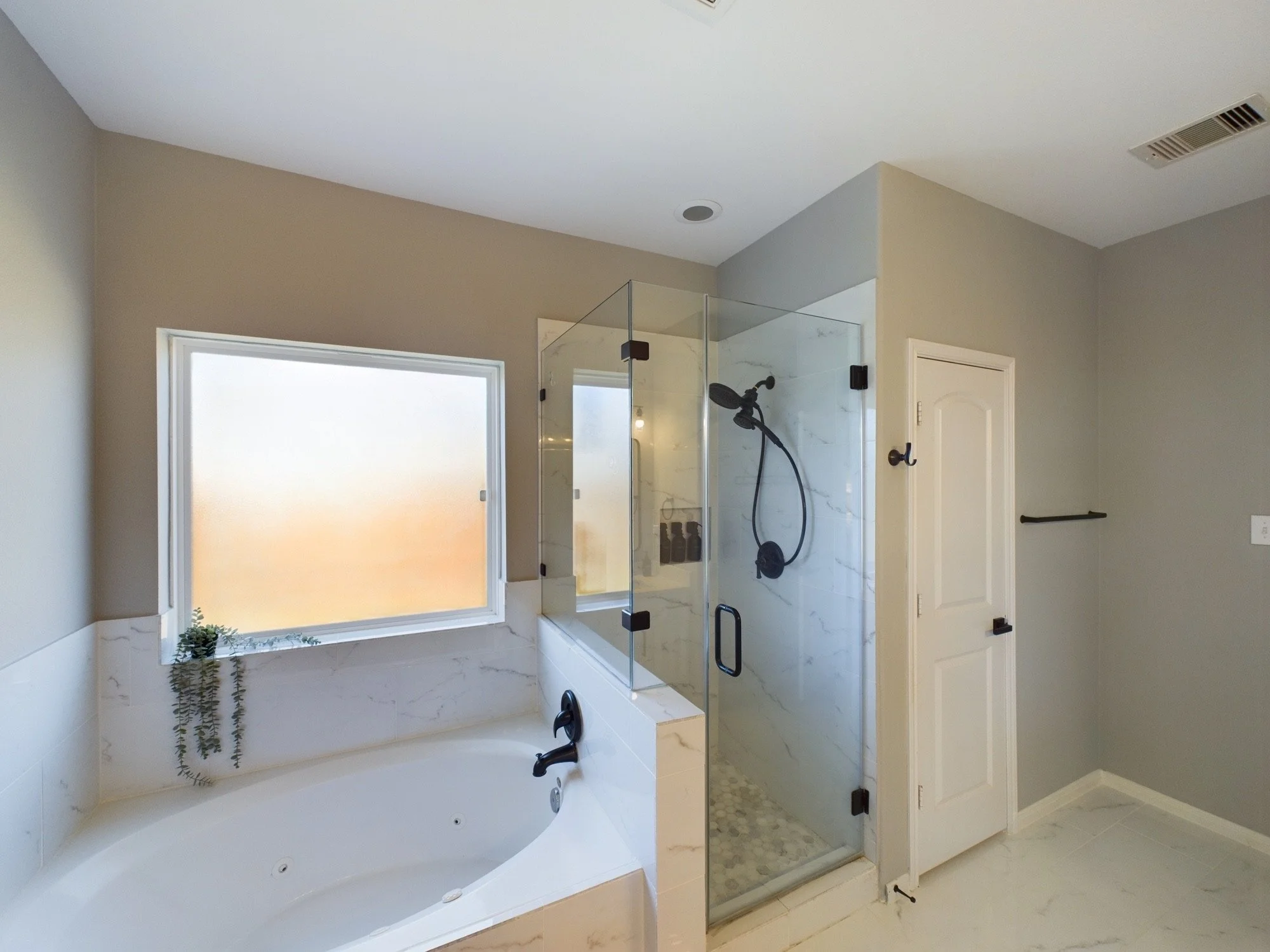 A modern bathroom features a large bathtub, a glass shower enclosure, a window, and light-colored walls.