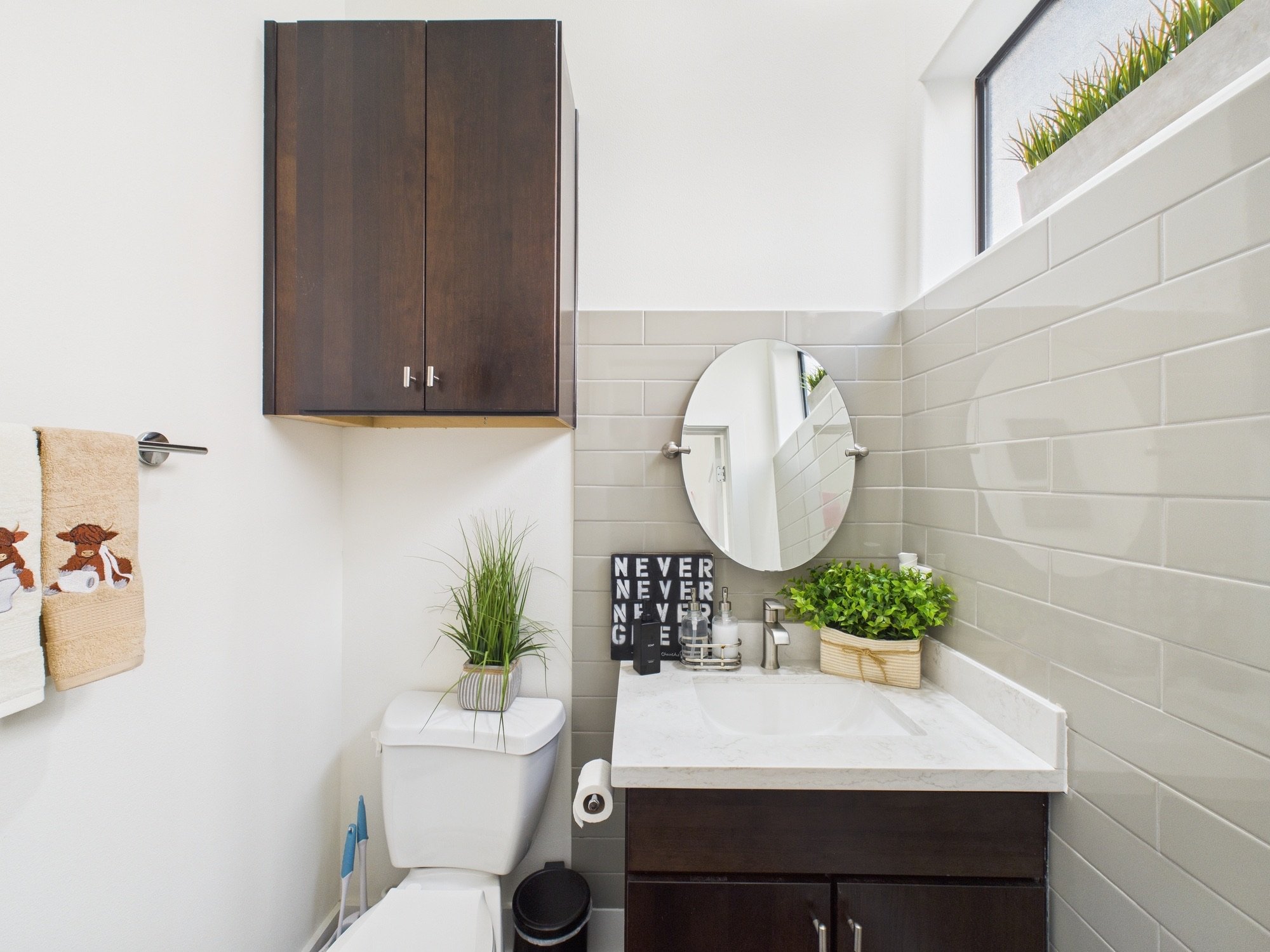Small bathroom with white walls and beige tiles, dark wood cabinet above toilet, round mirror on wall, black sign with white letters 'NEVER' stacked, green plants on counter, hand towels hanging on towel bar, window with plants outside.