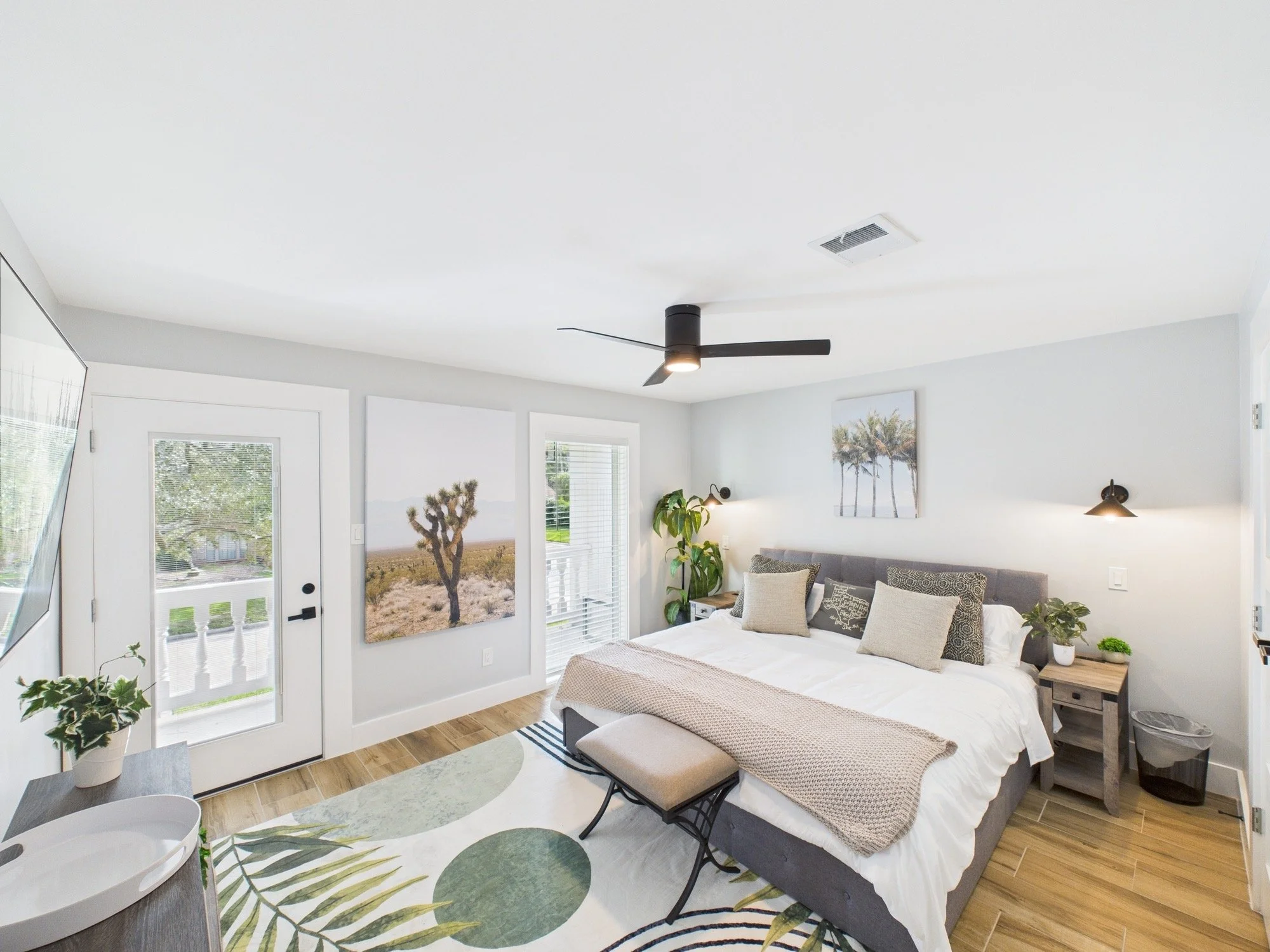 Bright bedroom with white walls, ceiling fan, artwork of palm trees, and large windows showing desert landscape outside.