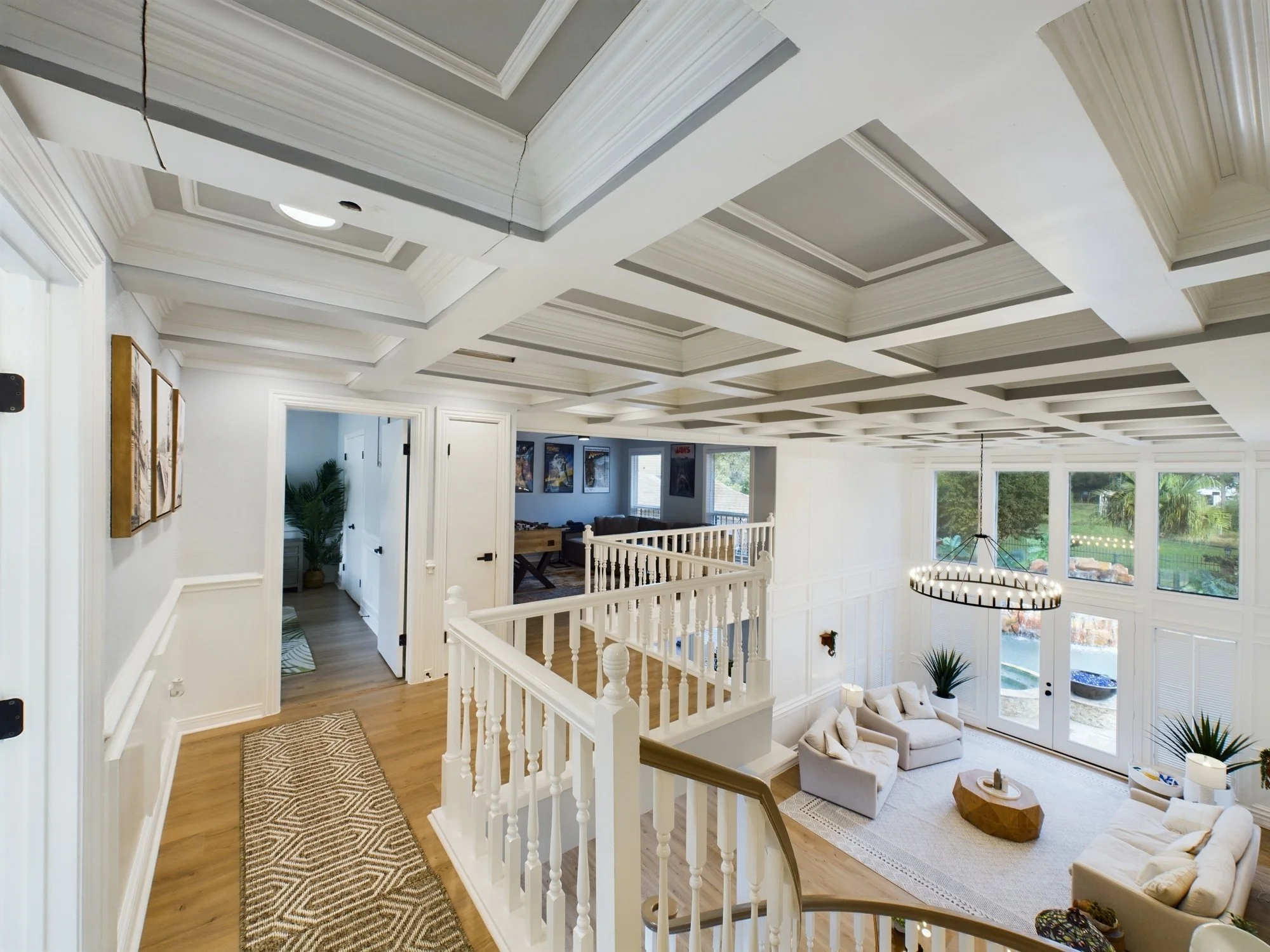 Interior view of a house with a two-story ceiling, white walls, large windows, and a living area with a cream sofa, wooden coffee table, and decorative plants.