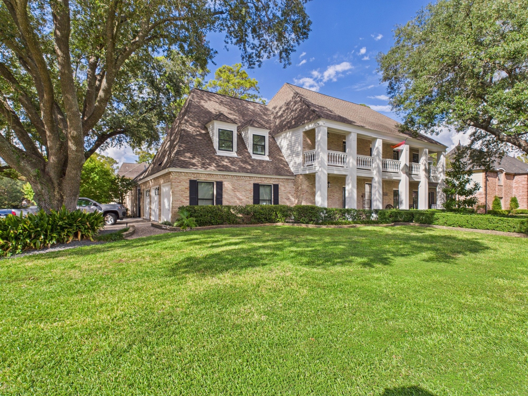 View of a large brick house with white columns on a porch, surrounded by green lawns and trees, under a blue sky with clouds.