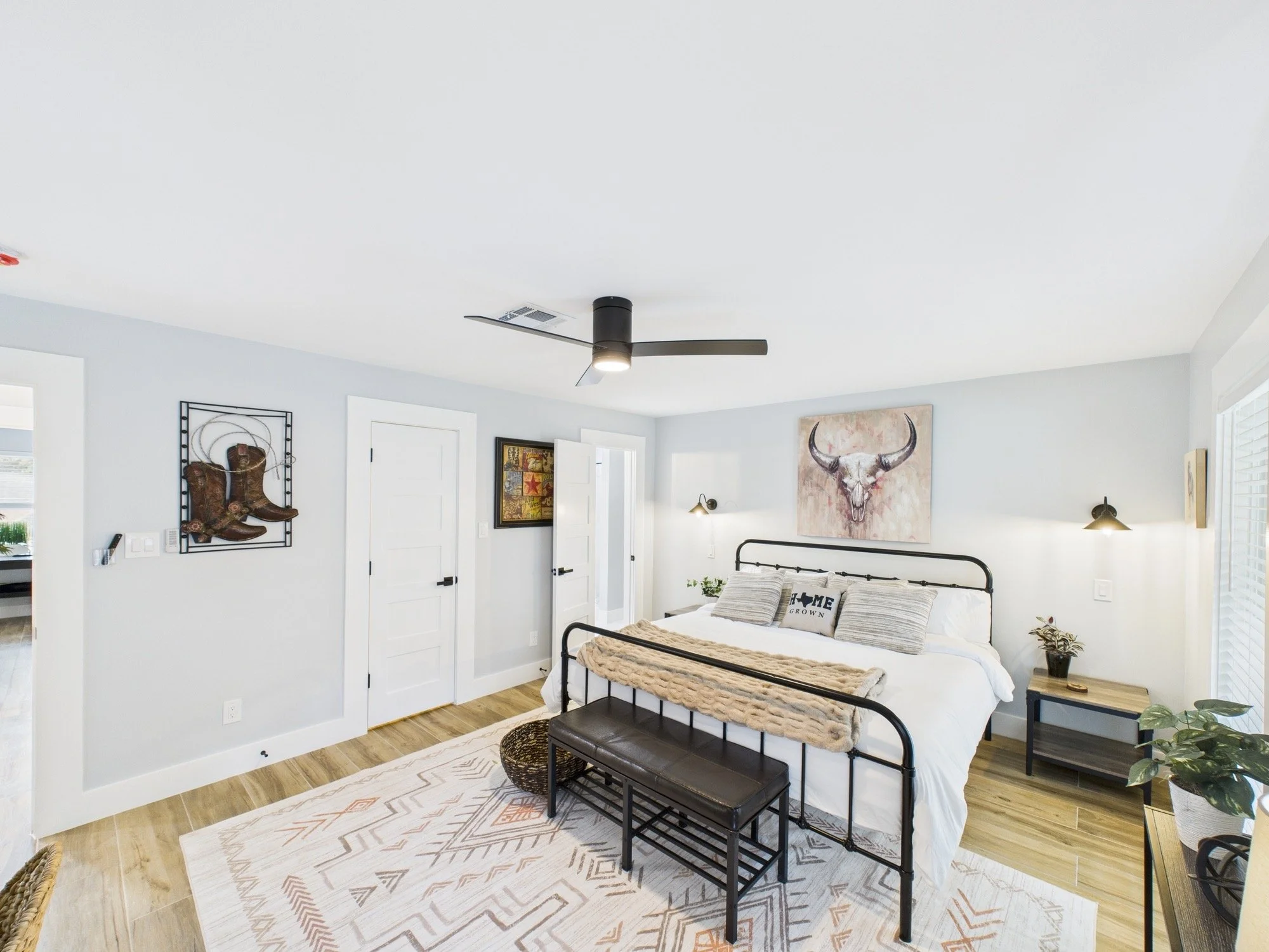 A bedroom with a metal bed frame, a cozy blanket, and decorative pillows that say 'Home Grown.' Artwork on the walls includes a bull skull painting and cowboy boots sculpture. There is a white door and a window with blinds, with plants and lamps arou