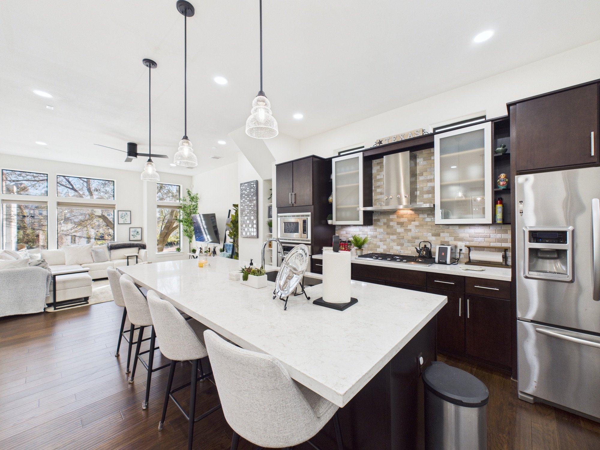 Modern open-concept kitchen and living room with white cabinetry, dark wood accents, pendant lights, large windows, and dining area with light fabric chairs.
