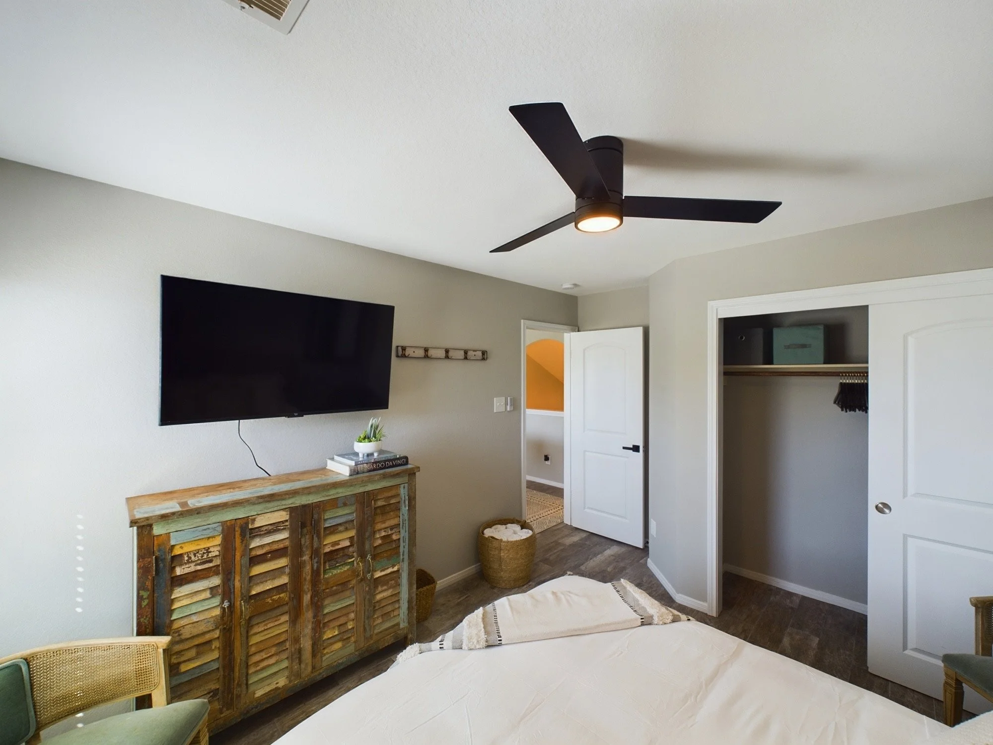 A bedroom with a ceiling fan, a wall-mounted TV, a rustic wooden dresser, a basket, and an open closet.