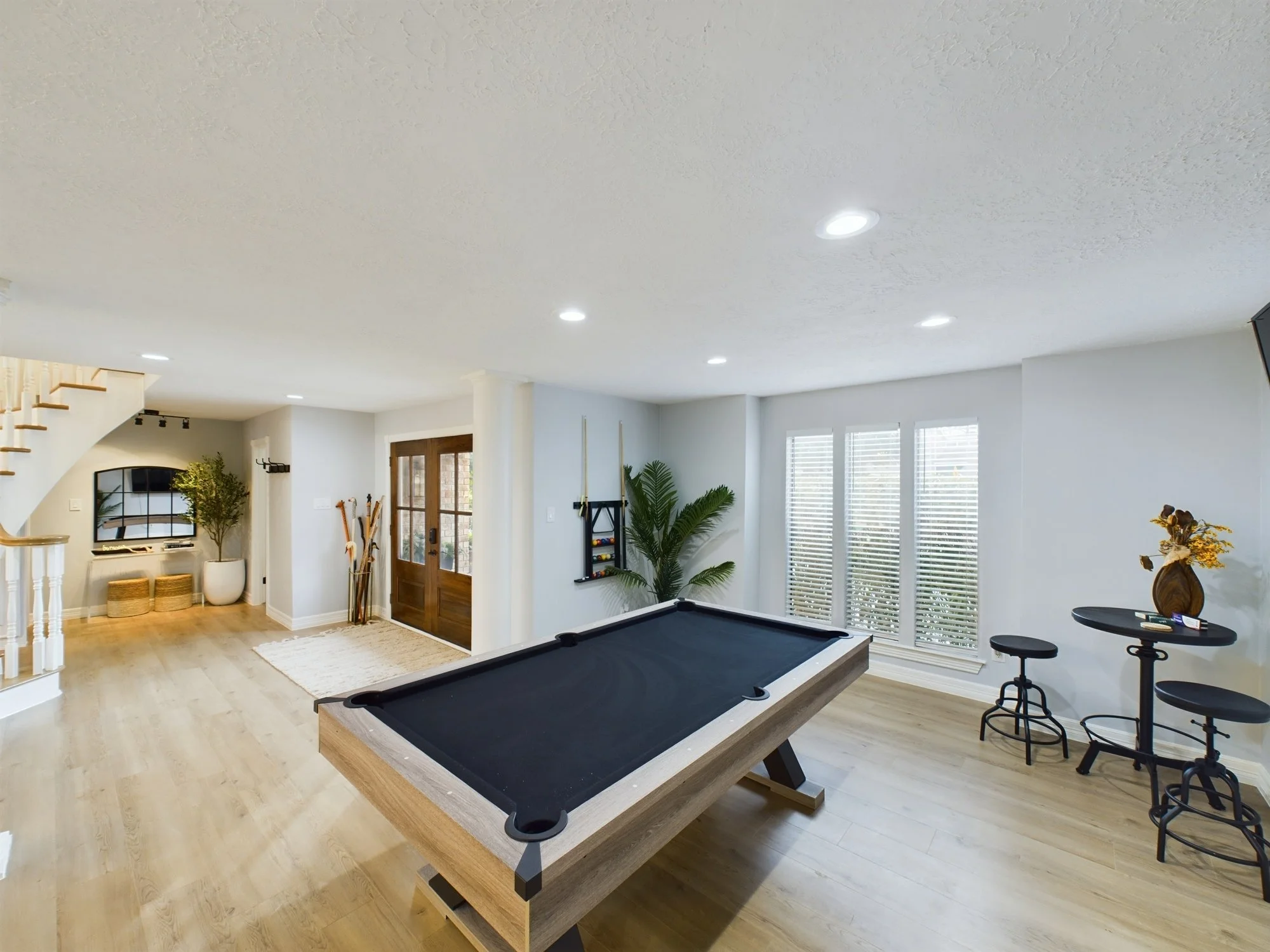 Living room with white walls, large windows with blinds, wooden floor, pool table in the center, plant near the windows, staircase on the left, TV and decorative items on the wall, bar stools on the right.