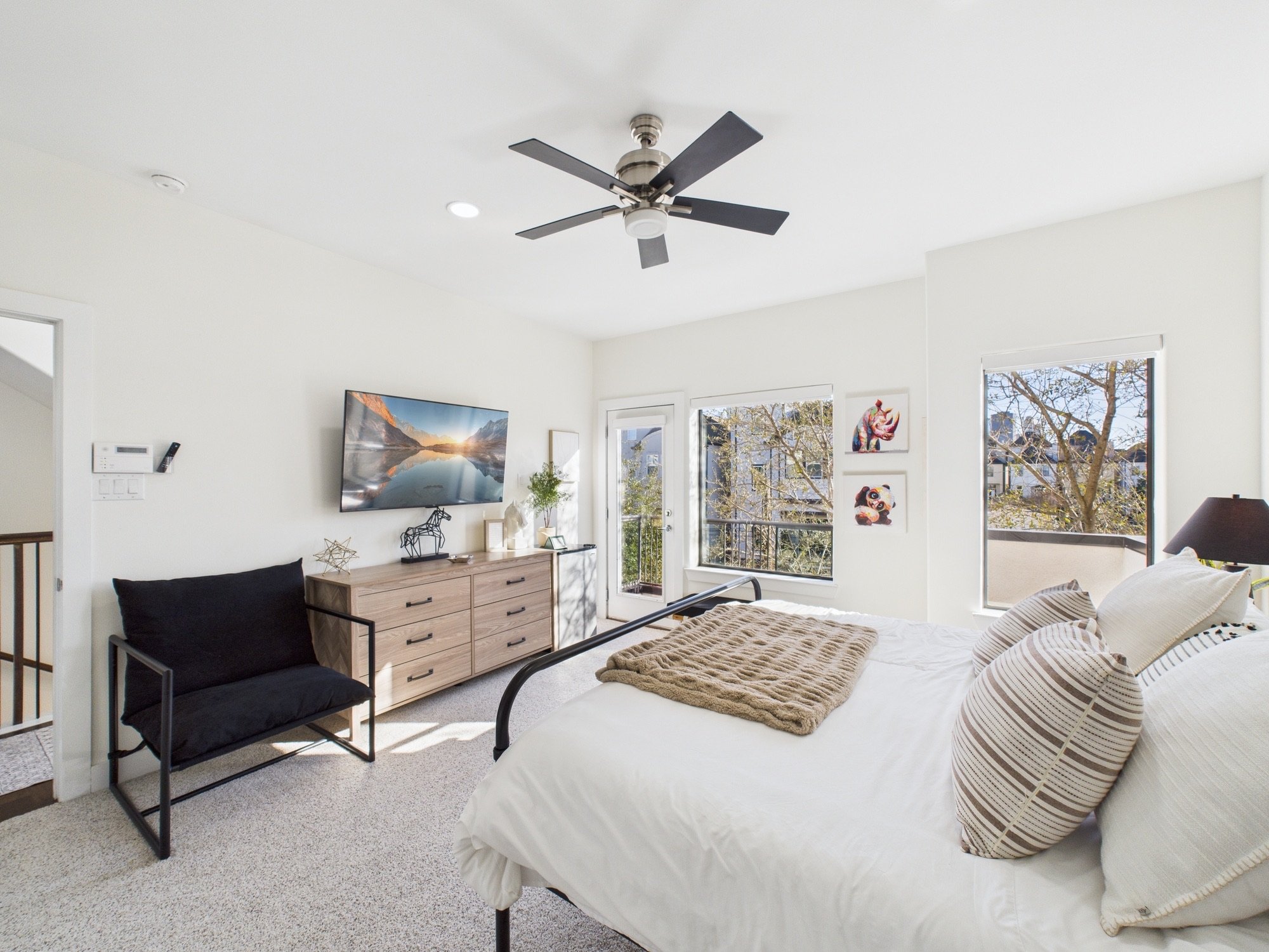 A bright bedroom featuring a bed with striped pillows and a furry blanket, a wooden dresser with decorative items, a wall-mounted TV, and large windows showing trees outside.
