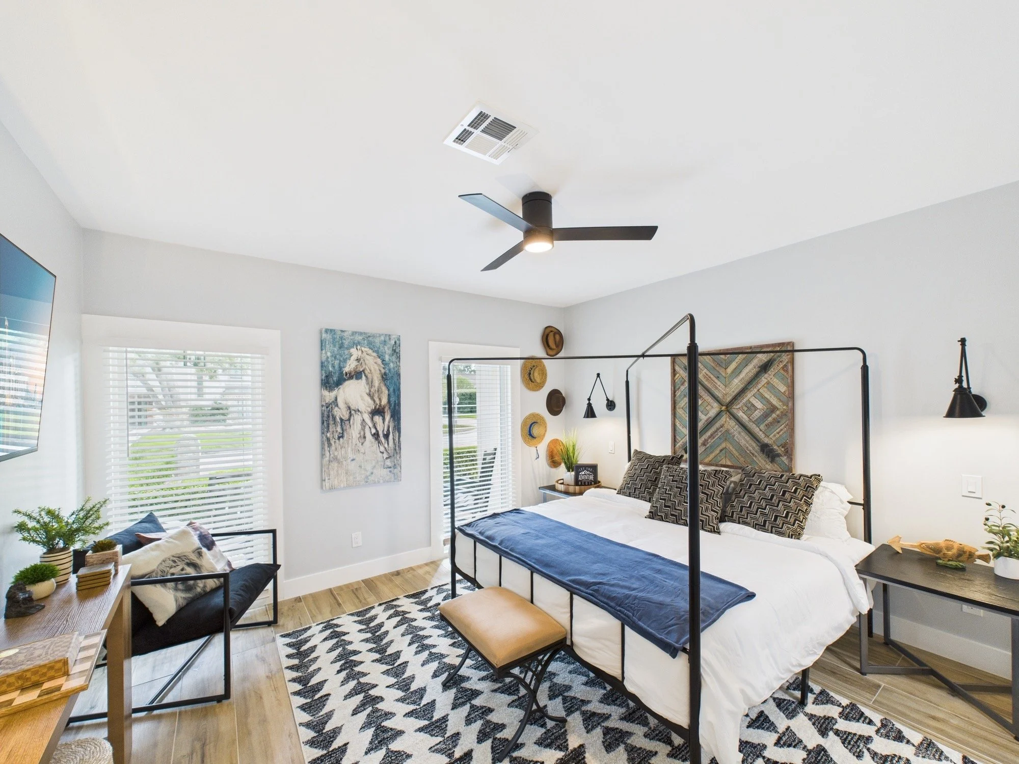 A modern bedroom with a bed and black metal frame, black and white chevron pillows, and a black and white geometric rug. There is a painting of a white horse on the wall, a small table with plants and decorative items, and a black table lamp. The roo