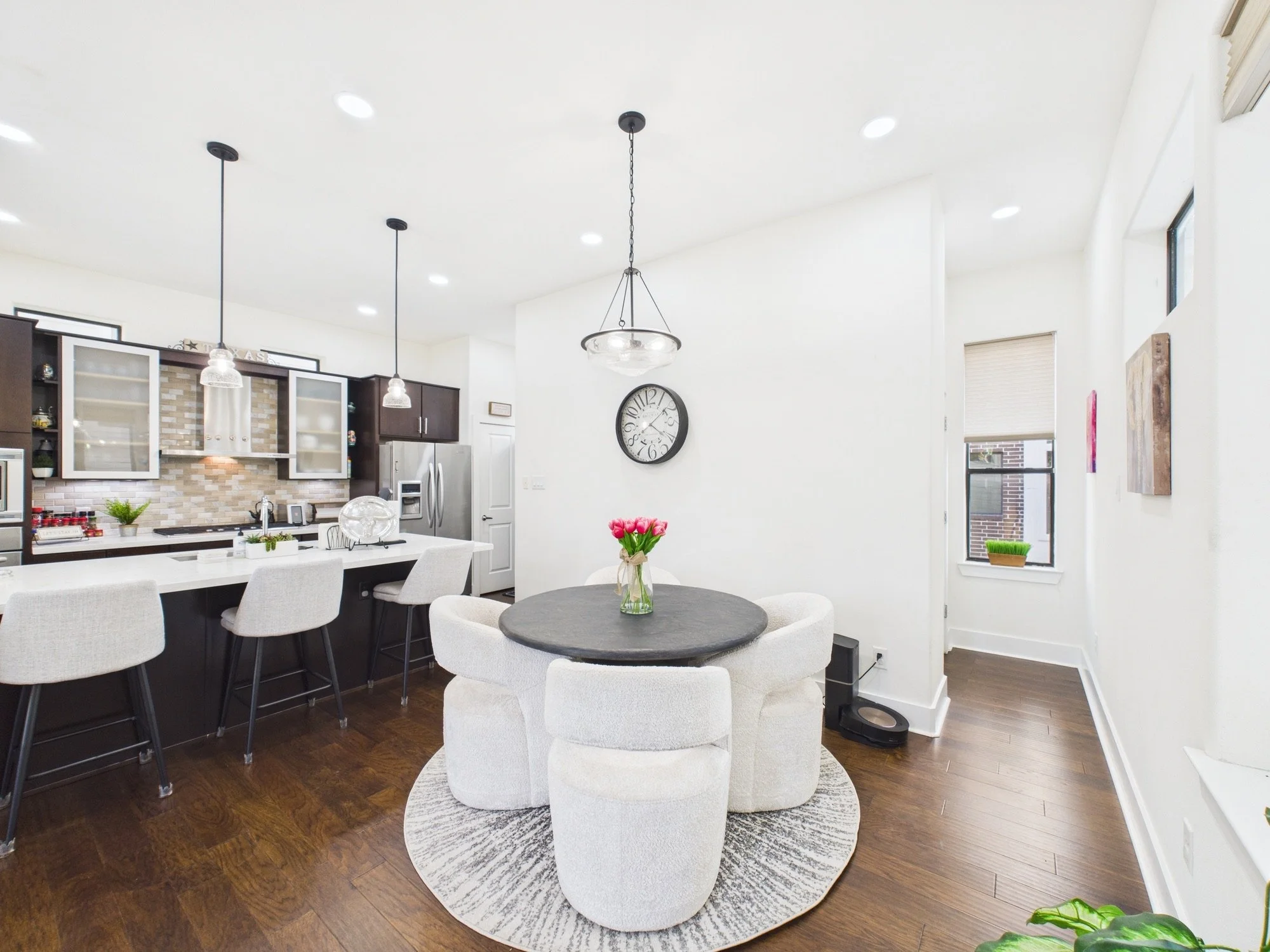Modern kitchen and dining area with white walls, dark wood flooring, a round dining table with white upholstered chairs, a small vase of pink tulips, a black round clock on the wall, and kitchen cabinetry with glass doors, stainless steel appliances,