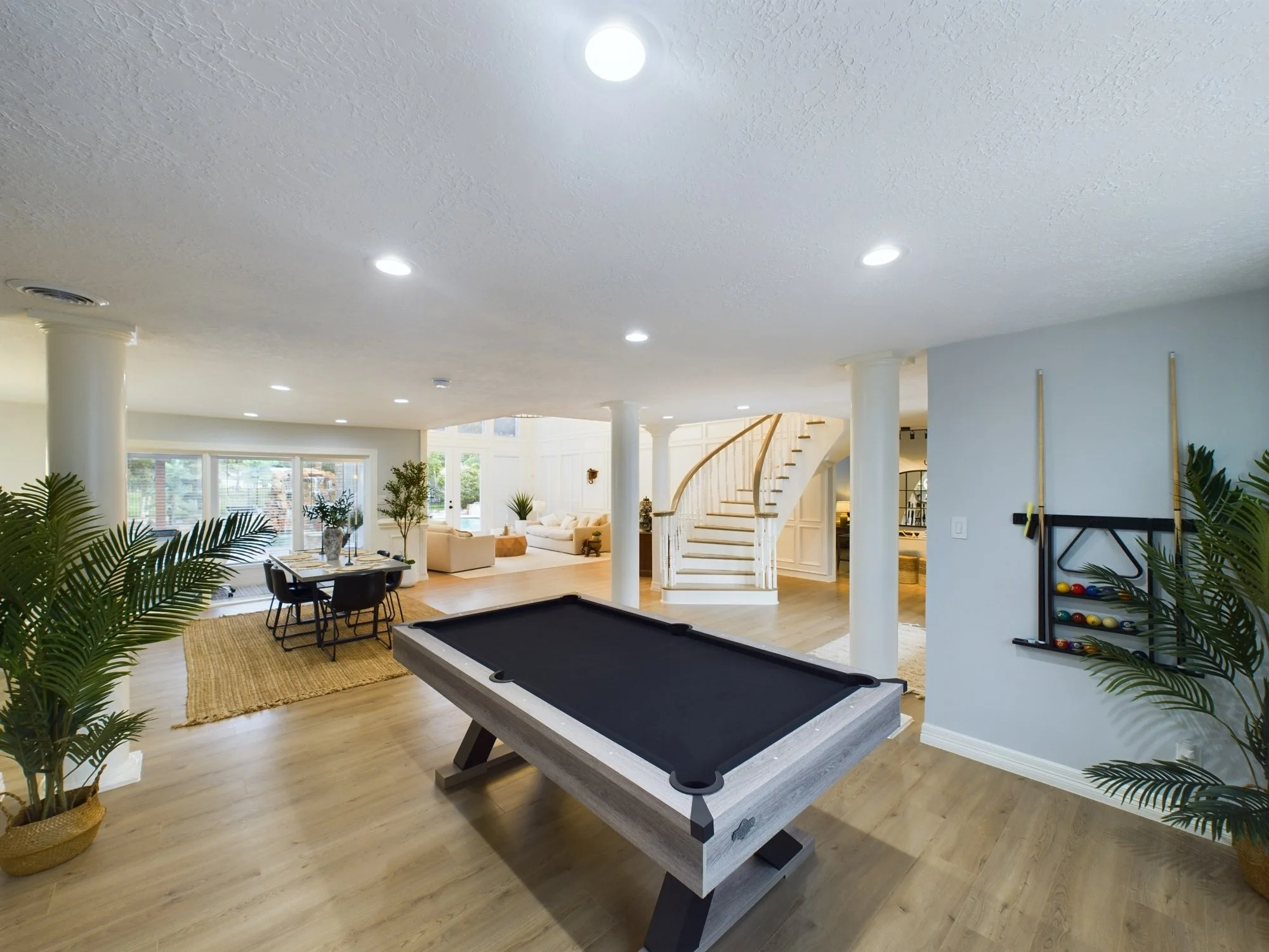 Living room with pool table, beige sofa, black dining table with chairs, houseplants, staircase, and large windows.