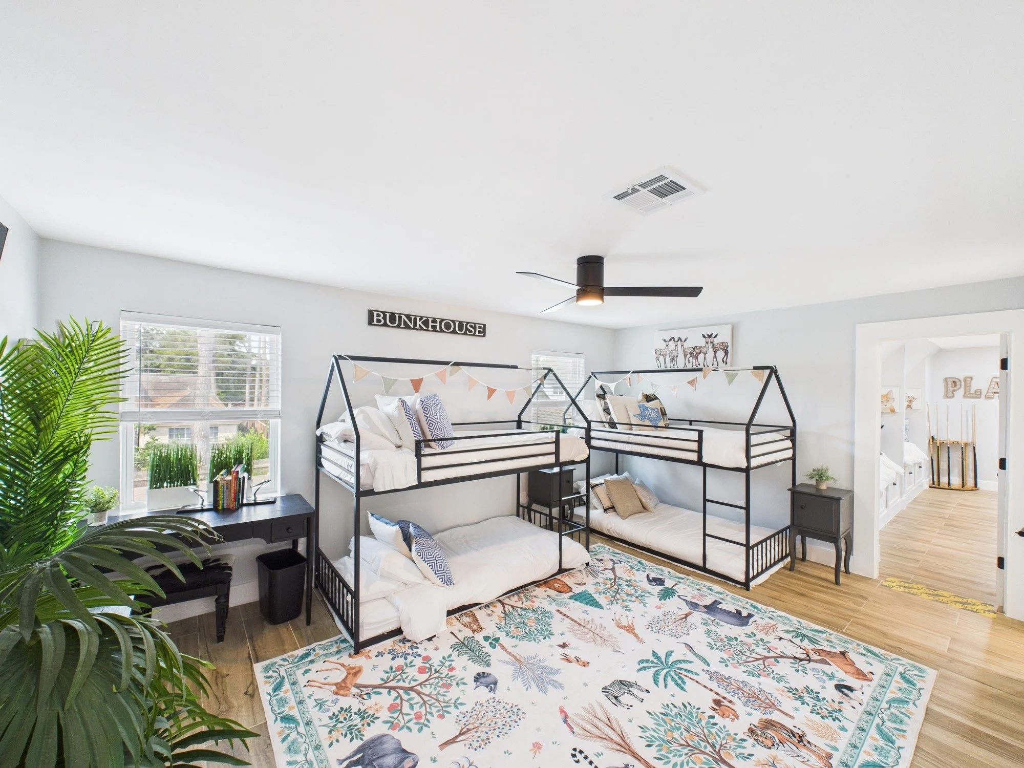 A bedroom with two bunk beds arranged in an L-shape, decorated with patterned pillows and a colorful animal-themed rug. There is a window with blinds, green plants, and a wall sign that reads 'BUNKHOUSE.'