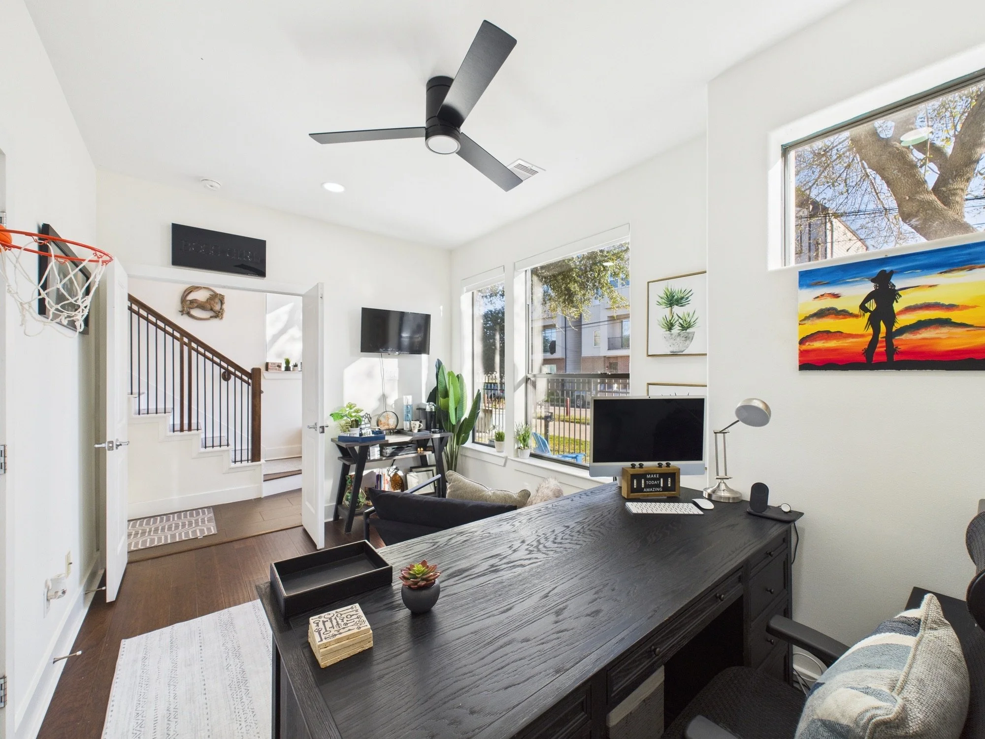 Bright living room with large windows, white walls, a modern black ceiling fan, artwork on walls, a TV, a dining table with decorative items, and a sports ball hoop on the wall.