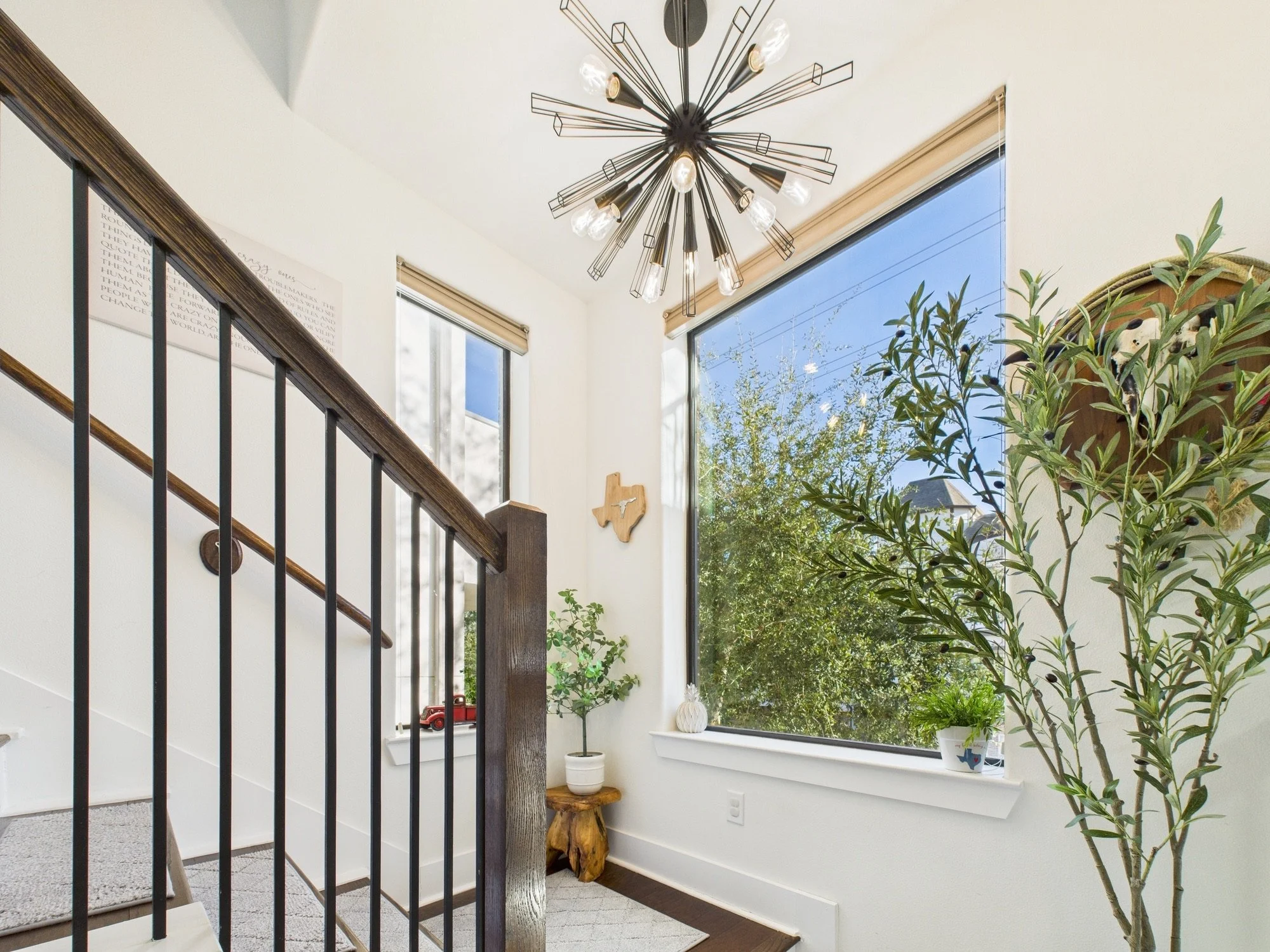 A bright living space with high ceilings, large windows, and a modern chandelier. There are potted plants, a wooden table with a white vase, and artwork on the wall. The space features a staircase with black railing.