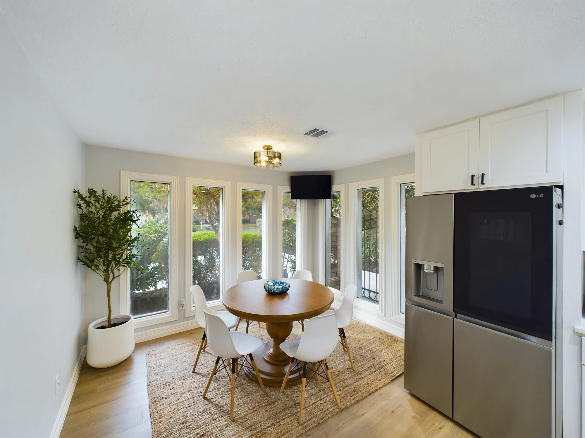 Modern dining area with a round wooden table surrounded by white chairs, large windows with trees outside, a potted plant, a ceiling light, and a wall-mounted TV.