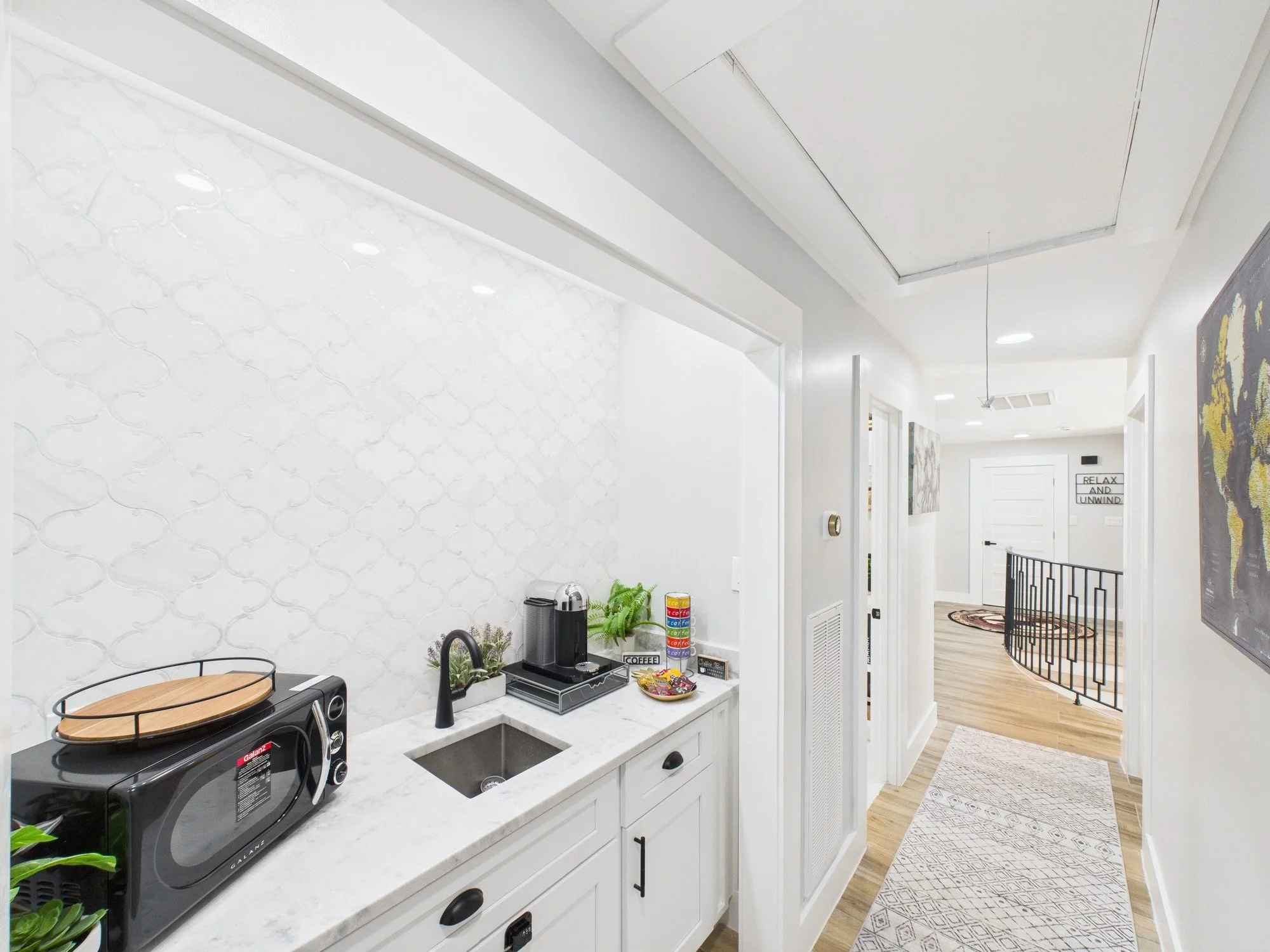 White kitchen counter with coffee machine, microwave, and small kitchen accessories. Bright wall with patterned backsplash. Hallway with wooden flooring, artwork, and a black railing.