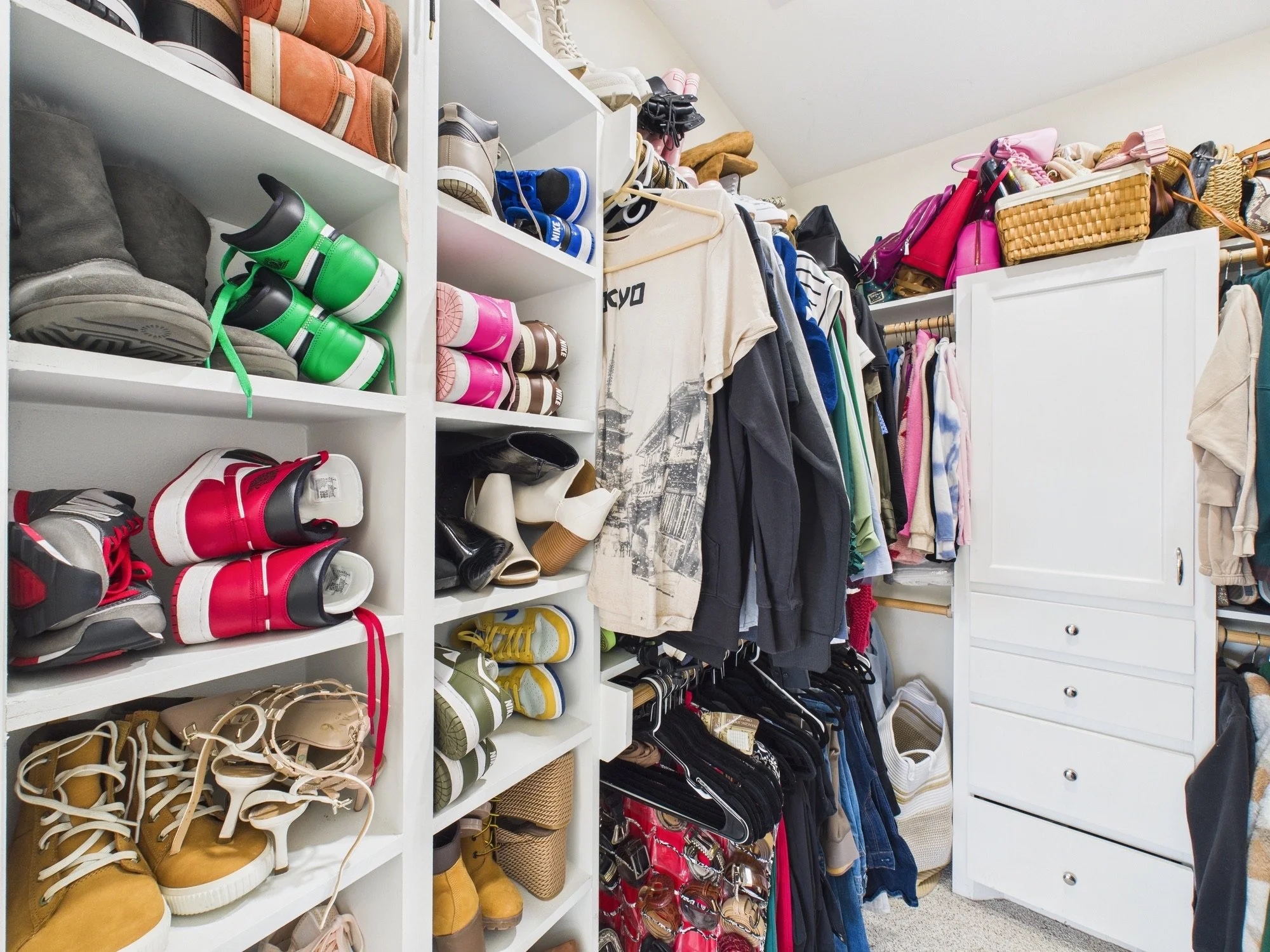 A walk-in closet with white shelves and drawers filled with shoes, clothing, and handbags. Shoes include sneakers, boots, sandals, and high heels, arranged on shelves. Clothing includes hanging shirts, dresses, and pants. Handbags are placed on the t