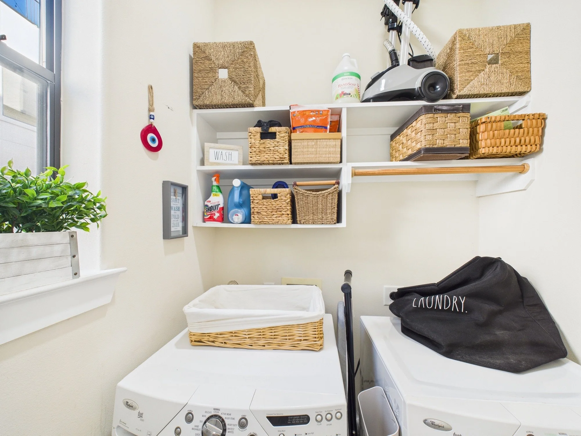 A laundry room with shelving holding baskets, cleaning supplies, and a vacuum. A washing machine and dryer are visible. There is a laundry bag on top of the dryer.