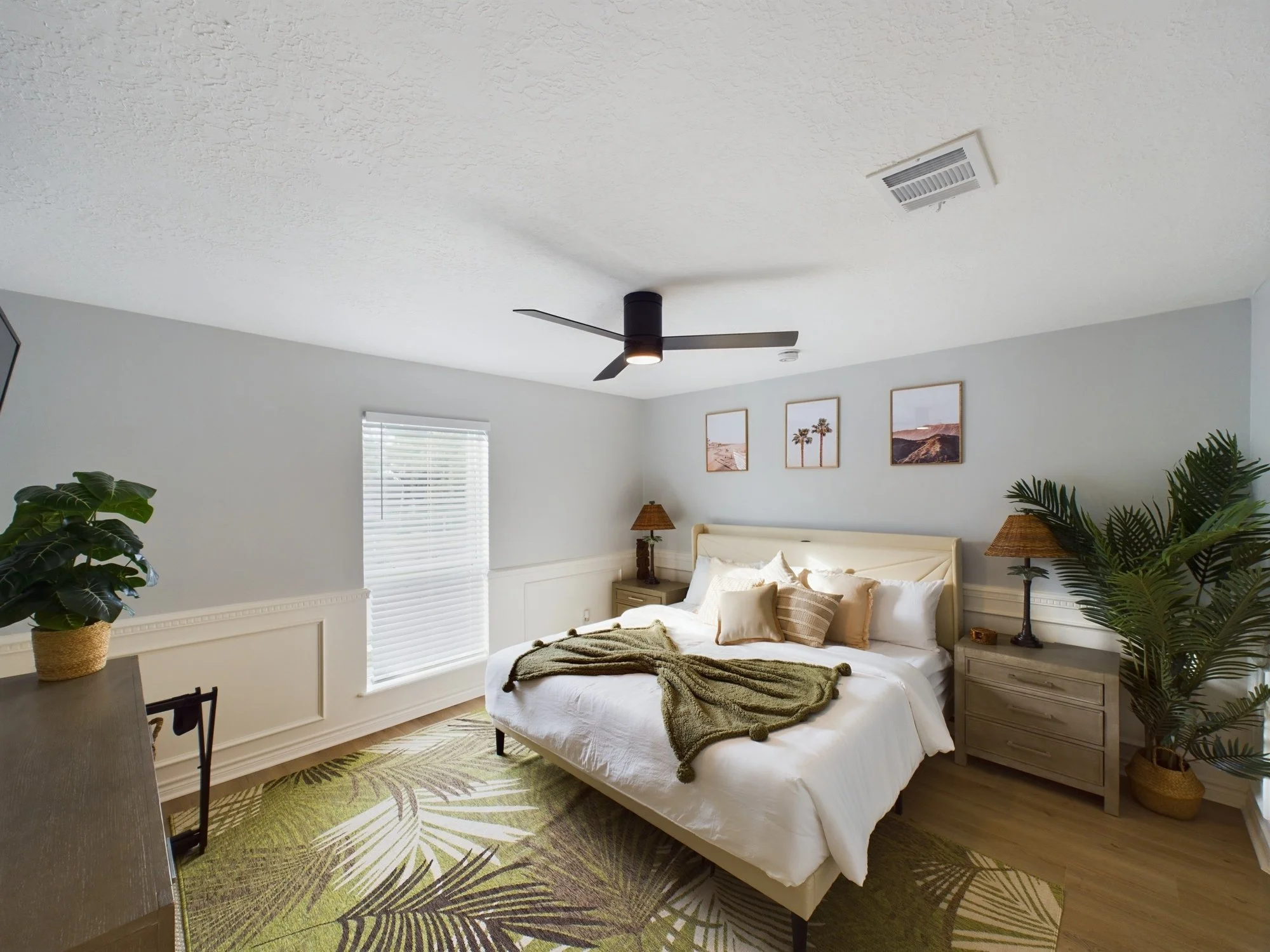 A bedroom with a white bed, beige headboard, and bedspread. Nightstands with lamps on each side, framed photographs of desert and palm trees hanging above. Green plants in pots, a window with blinds, and a patterned rug on hardwood floor. A ceiling f