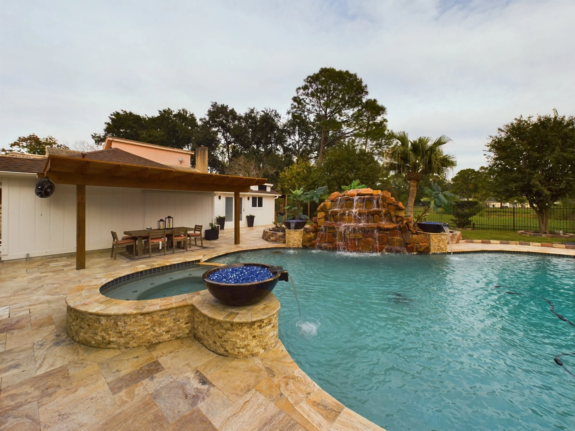 Backyard swimming pool with waterfall feature, outdoor dining area, and lush greenery with trees in the background.