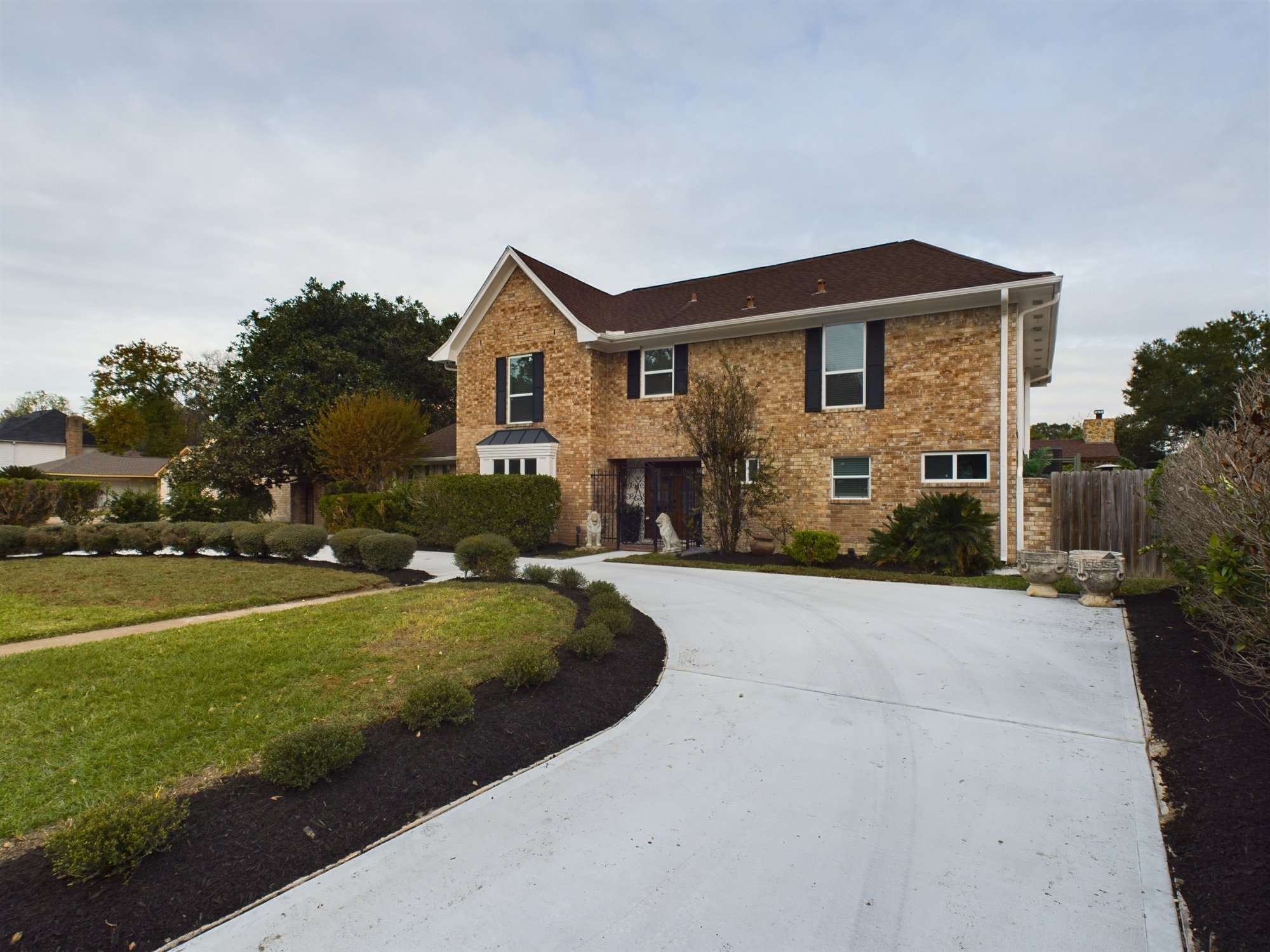 A two-story brick house with a brown roof and black shutters. It has a white fenced front yard with a pathway leading to the entrance, flanked by decorative lion statues and various shrubs. The sky is partly cloudy, and neighboring houses are visible