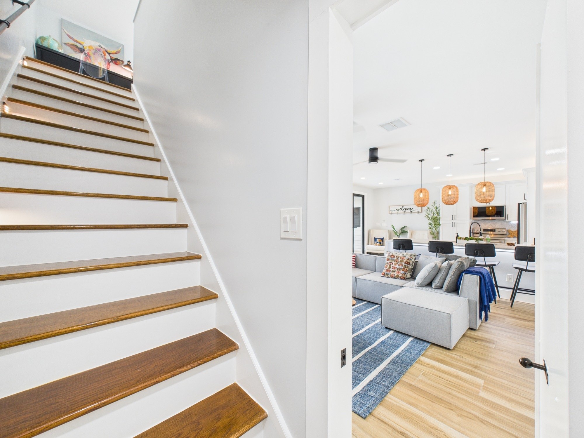 Interior of a modern home with a staircase leading to the upper floor and a living area visible through a doorway. The living room has a gray sectional sofa, a blue area rug, pendant lights, and various decorative items.