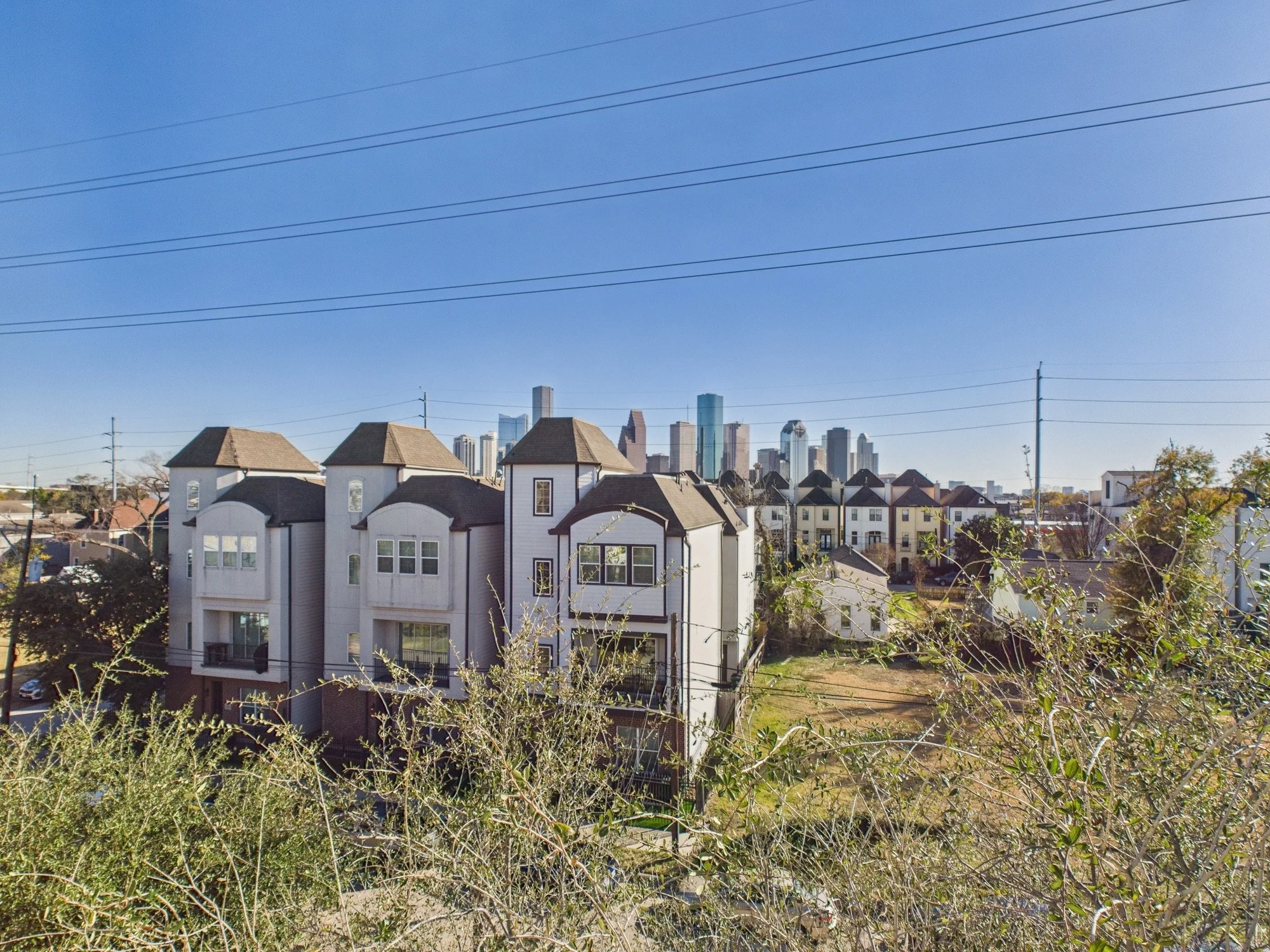 Residential neighborhood with multi-story white houses in the foreground and a city skyscraper skyline in the background, under a clear blue sky with utility power lines crossing the scene.