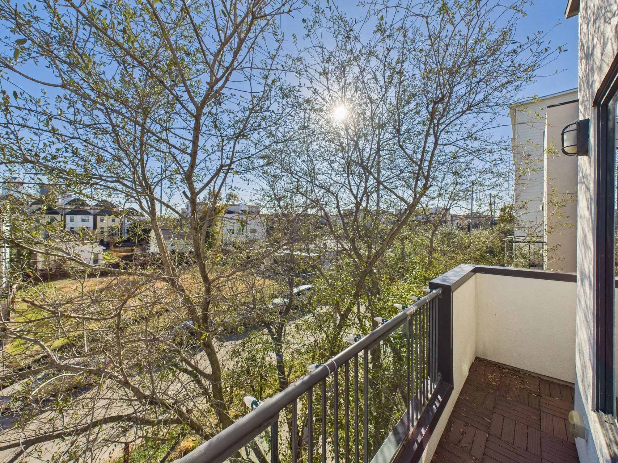 View from a balcony showing leafless tree branches illuminated by sunlight against a clear blue sky, with nearby buildings in the background.
