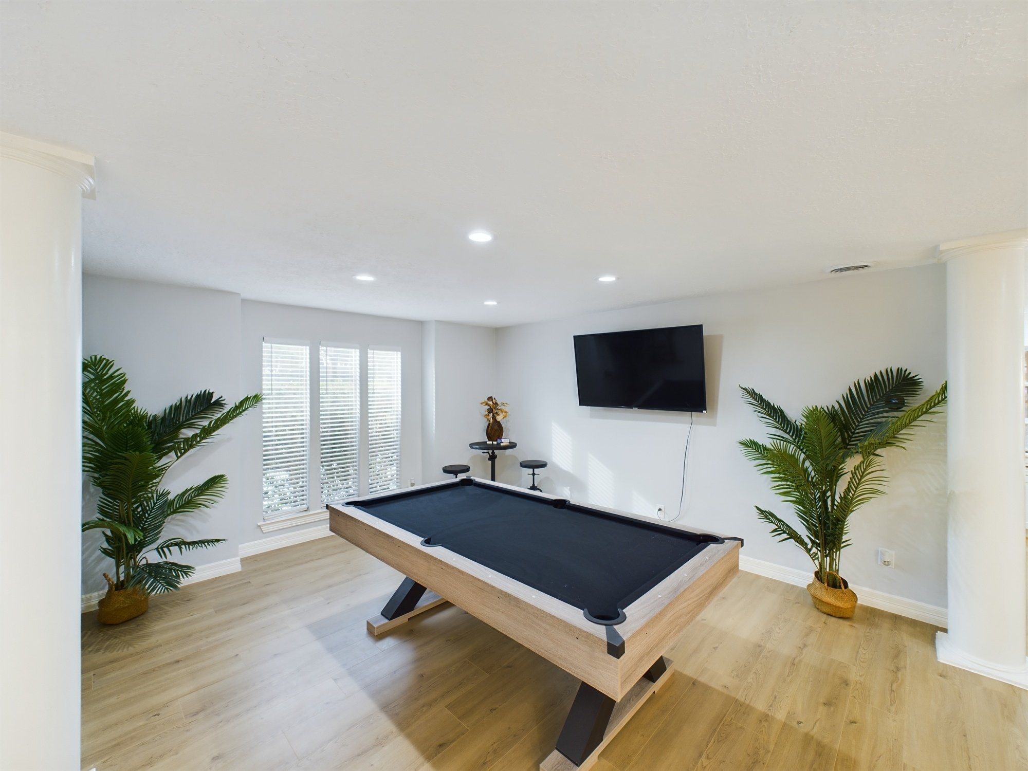 A living room with a pool table in the center, surrounded by two potted palm plants, a television mounted on the wall, and windows with white blinds.