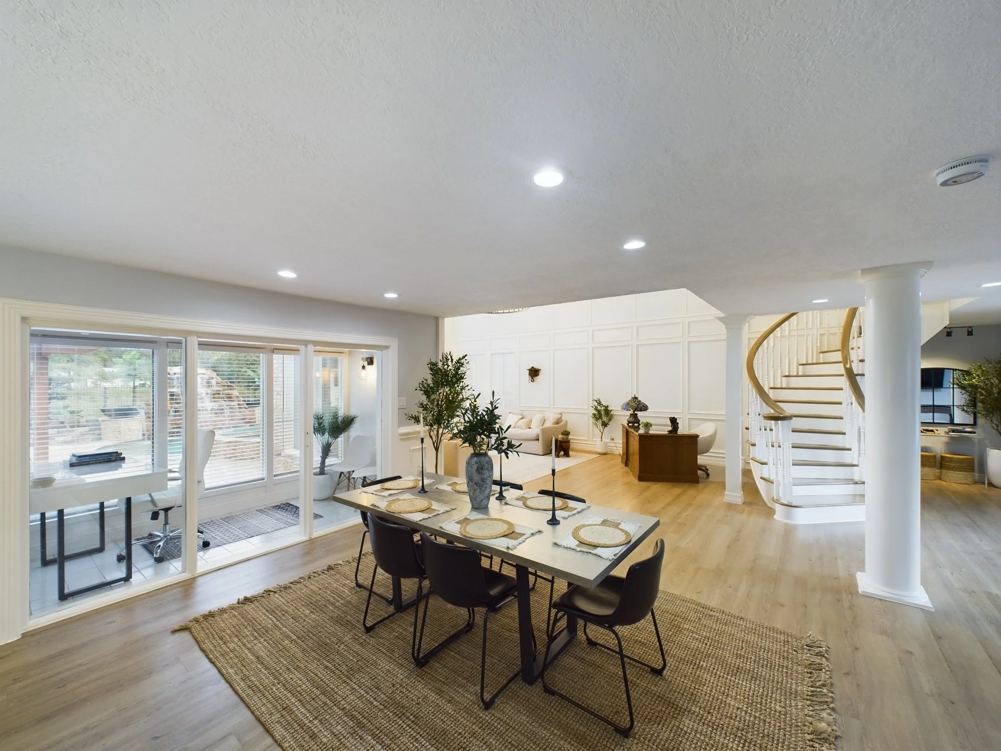Interior view of a spacious, well-lit living and dining area with large windows, a glass dining table with six chairs, and a staircase with a curved wooden banister. There are various decorative plants and a woven rug on the hardwood floor.