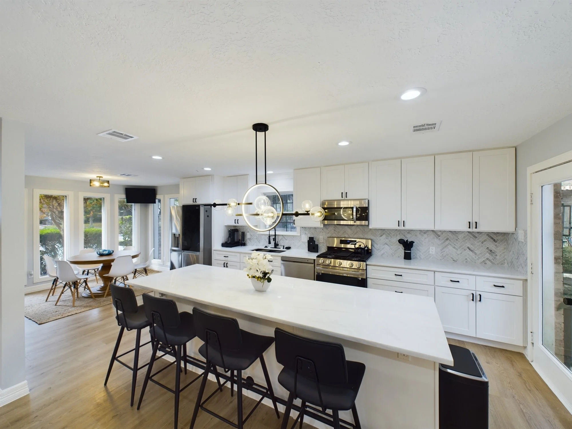 Modern kitchen with white cabinets, stainless steel appliances, island with black chairs, black pendant light, and dining area with light wooden floors and windows.