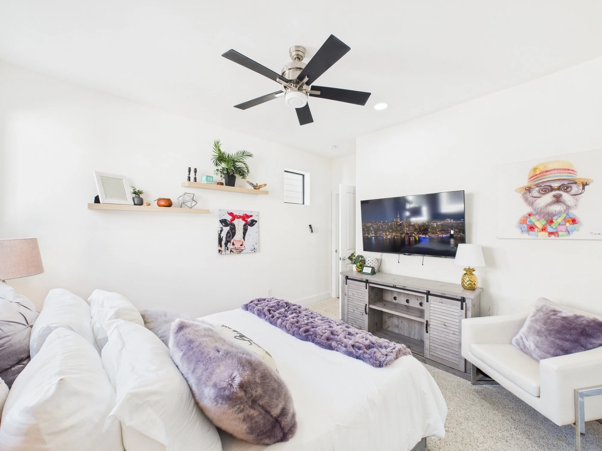 Living room with white walls, a large white sofa with purple faux fur pillows, a TV displaying a cityscape at night, and decorations including plants, artwork, and a gold pineapple-shaped lamp.