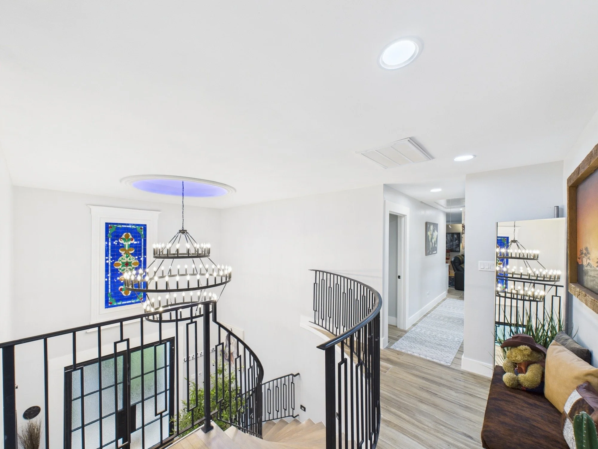 Interior view of a multi-story home with white walls, hardwood floors, a curved black staircase, a hanging chandelier with candles, two recessed ceiling lights, a stained glass window, and a sofa with teddy bears.