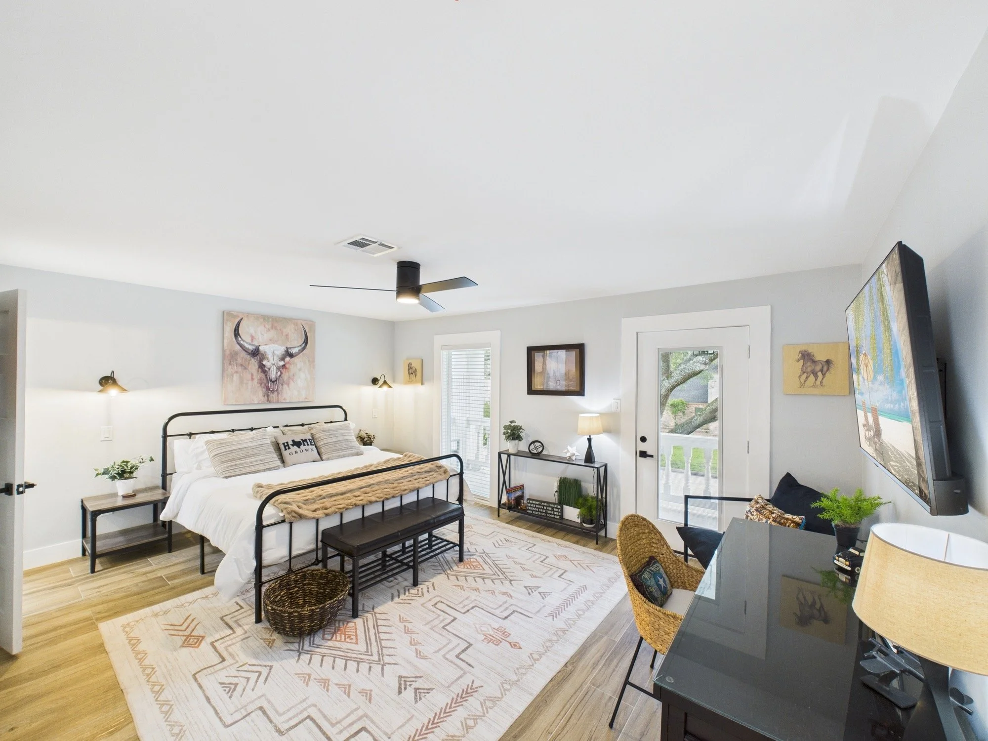 A bedroom with a bed featuring a black metal frame, white bedding, and decorative pillows. There is a small wooden nightstand with plants and books, a sitting area with wicker chairs, and wall art including a buffalo skull painting. Natural light fil