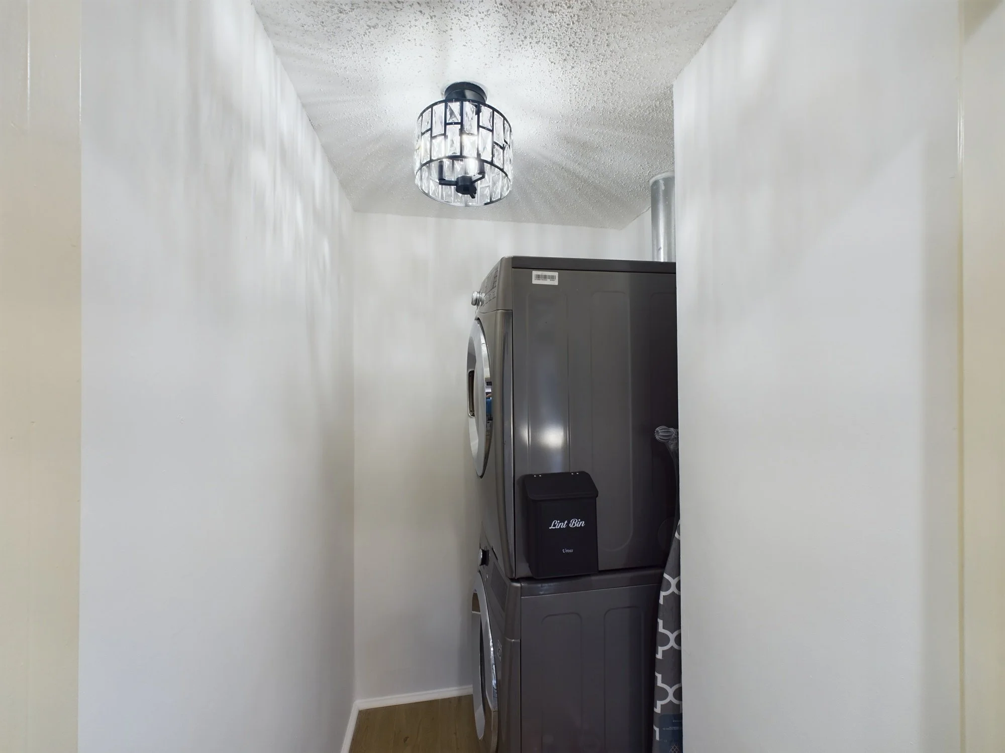 A laundry area with a stacked washer and dryer, a lint bin, plain white walls, and a ceiling light fixture.