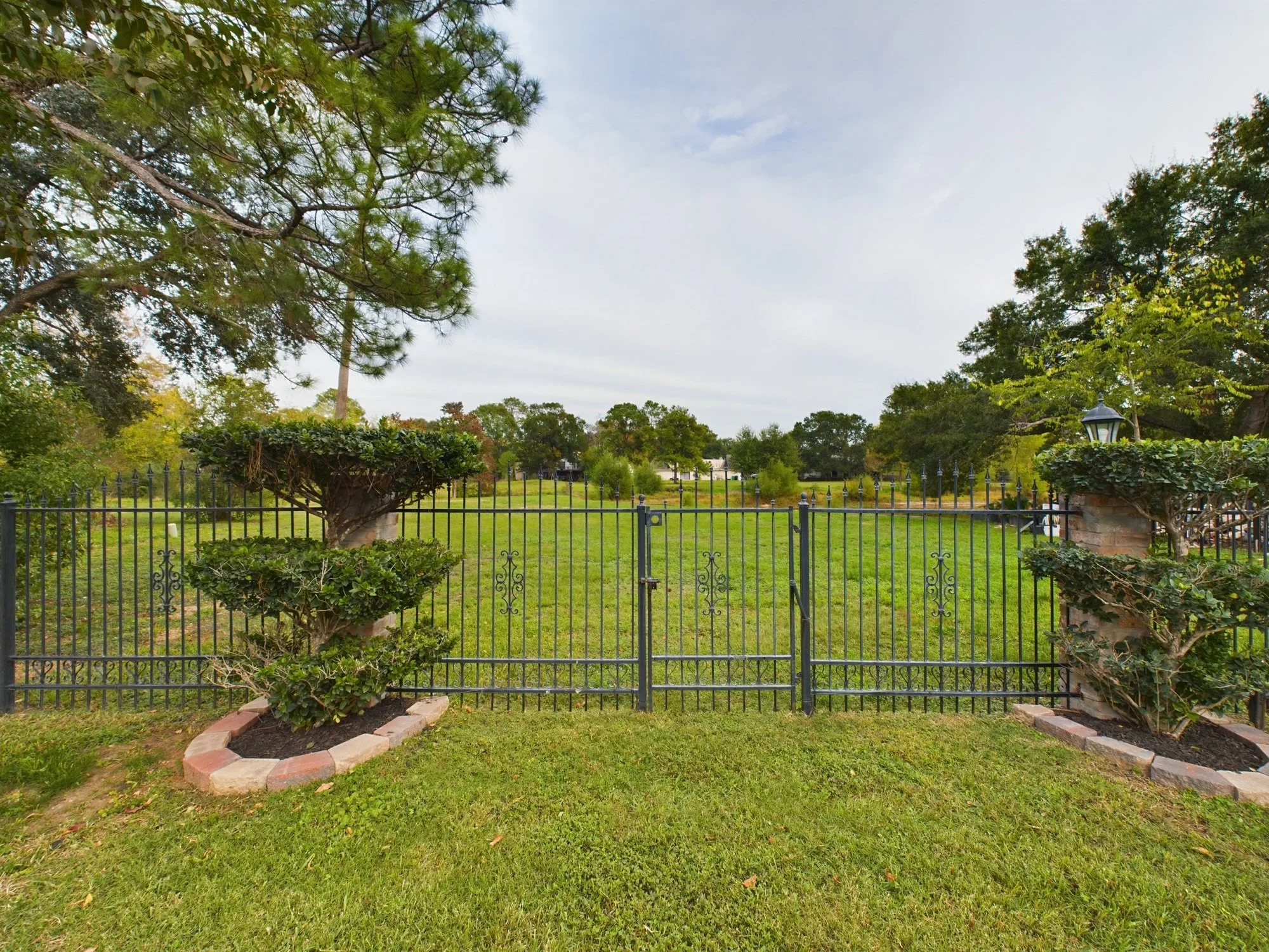 A view of a backyard with a black metal fence, with two sidewalk-edged flower beds with shrubs and a large tree on each side, one with arranged brick borders at the base, the other with a wrought iron fence and a vintage lamp post. In the background,
