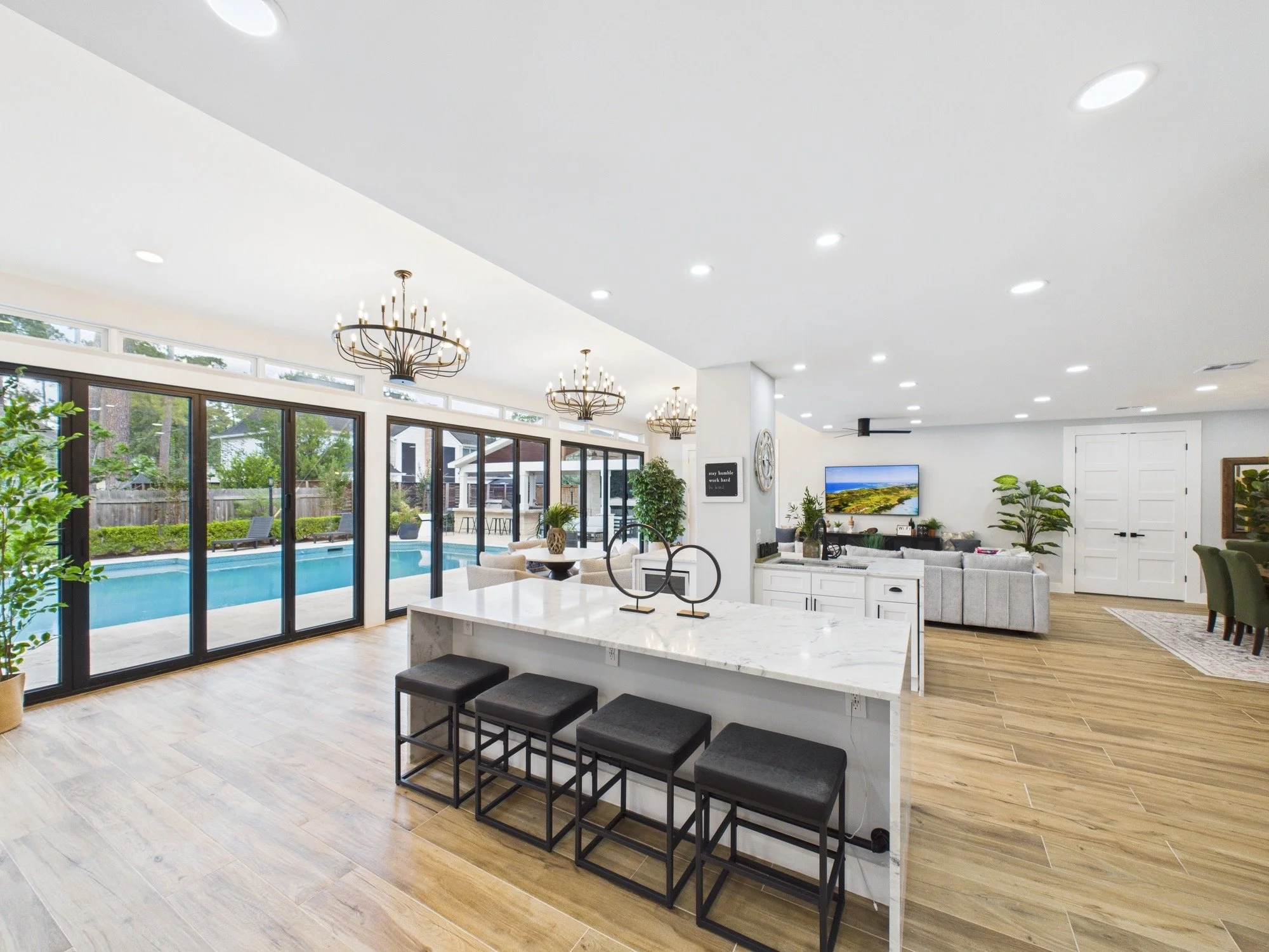 Interior view of a modern living room with large windows, a bedroom, a TV, plants, decor, and an open kitchen area with a kitchen island and bar stools.