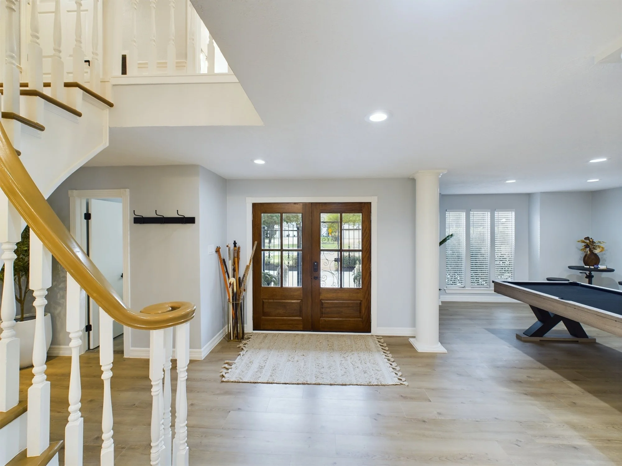 Interior view of a modern house foyer with a wooden front door, a beige rug on light wood flooring, and a staircase with white and gold railings on the left. There are coat hooks and a coat rack on the wall, with a bundle of walking sticks next to th