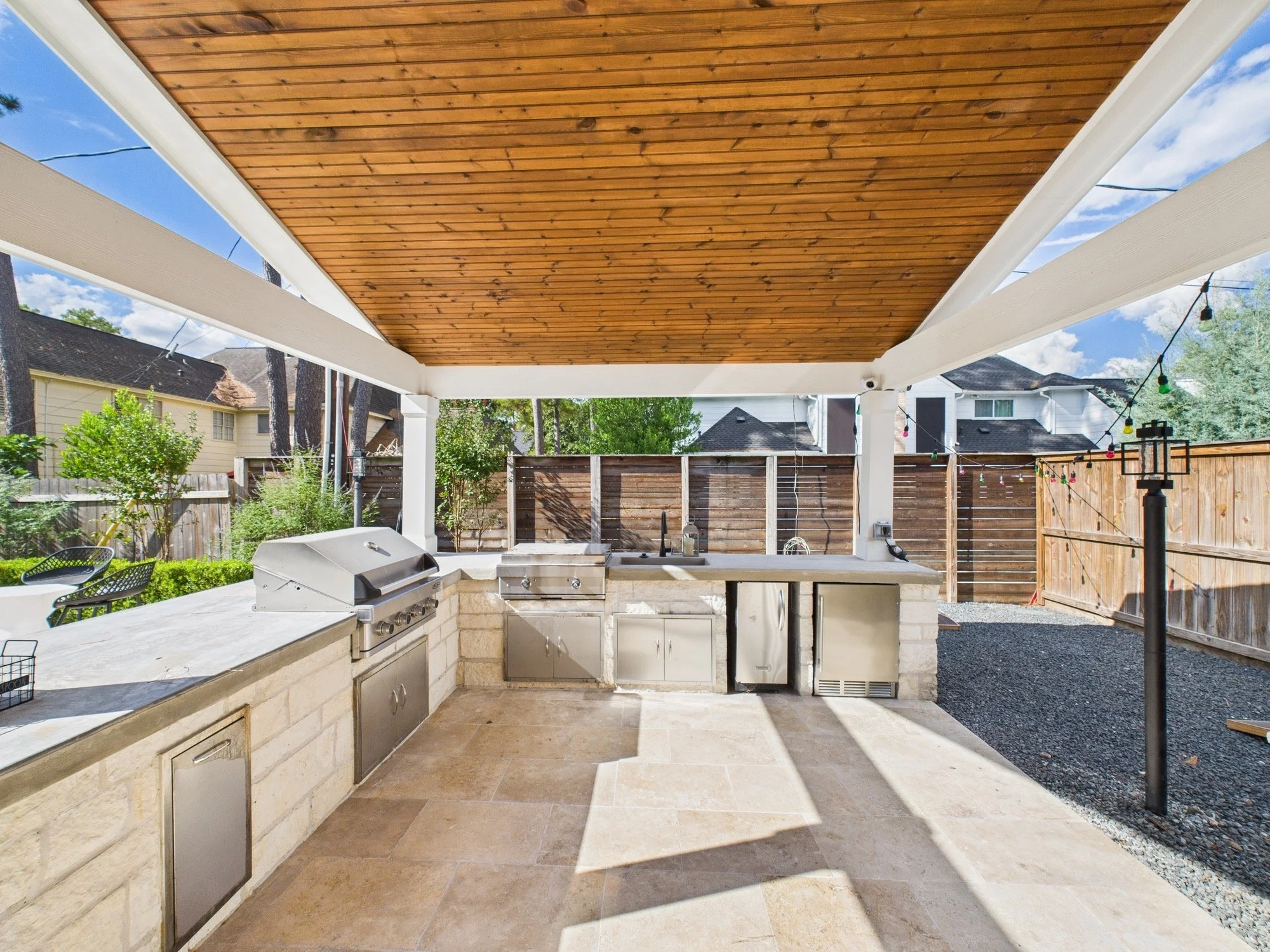 Outdoor kitchen with a stone counter, built-in grill, and a wooden ceiling under a covered patio, with a backyard enclosed by a wooden fence and neighboring houses visible in the background.