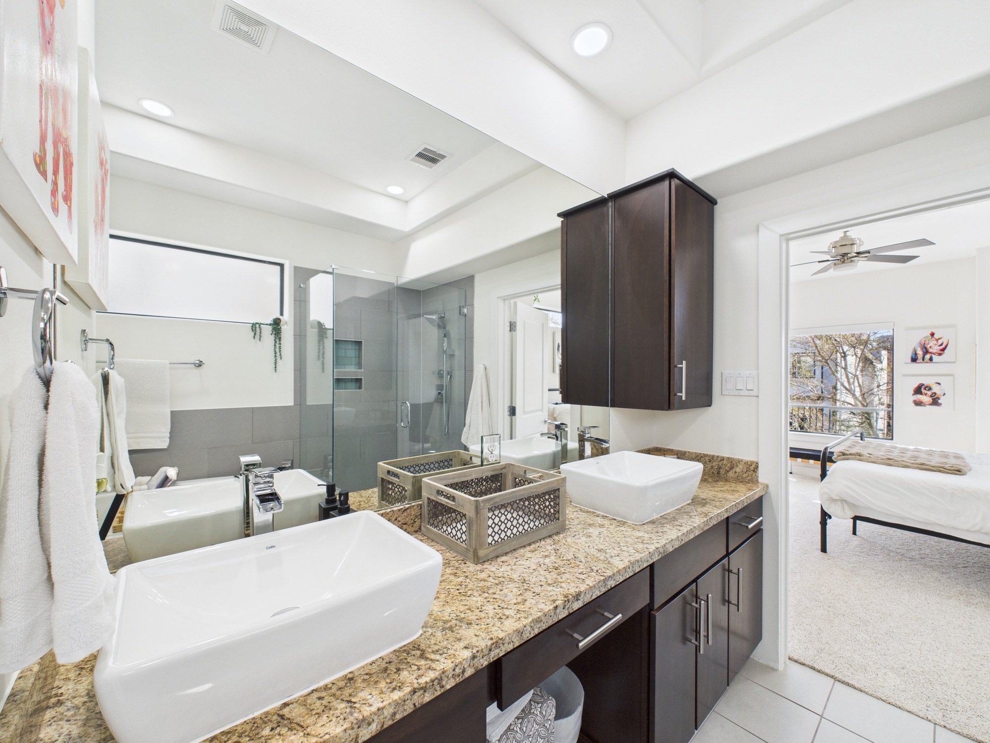 Modern bathroom with a double granite countertop, two vessel sinks, and a large mirror. Dark wood cabinets, a glass shower enclosure, and a window with a view of trees outside. Decorative artwork and small baskets on the counter.