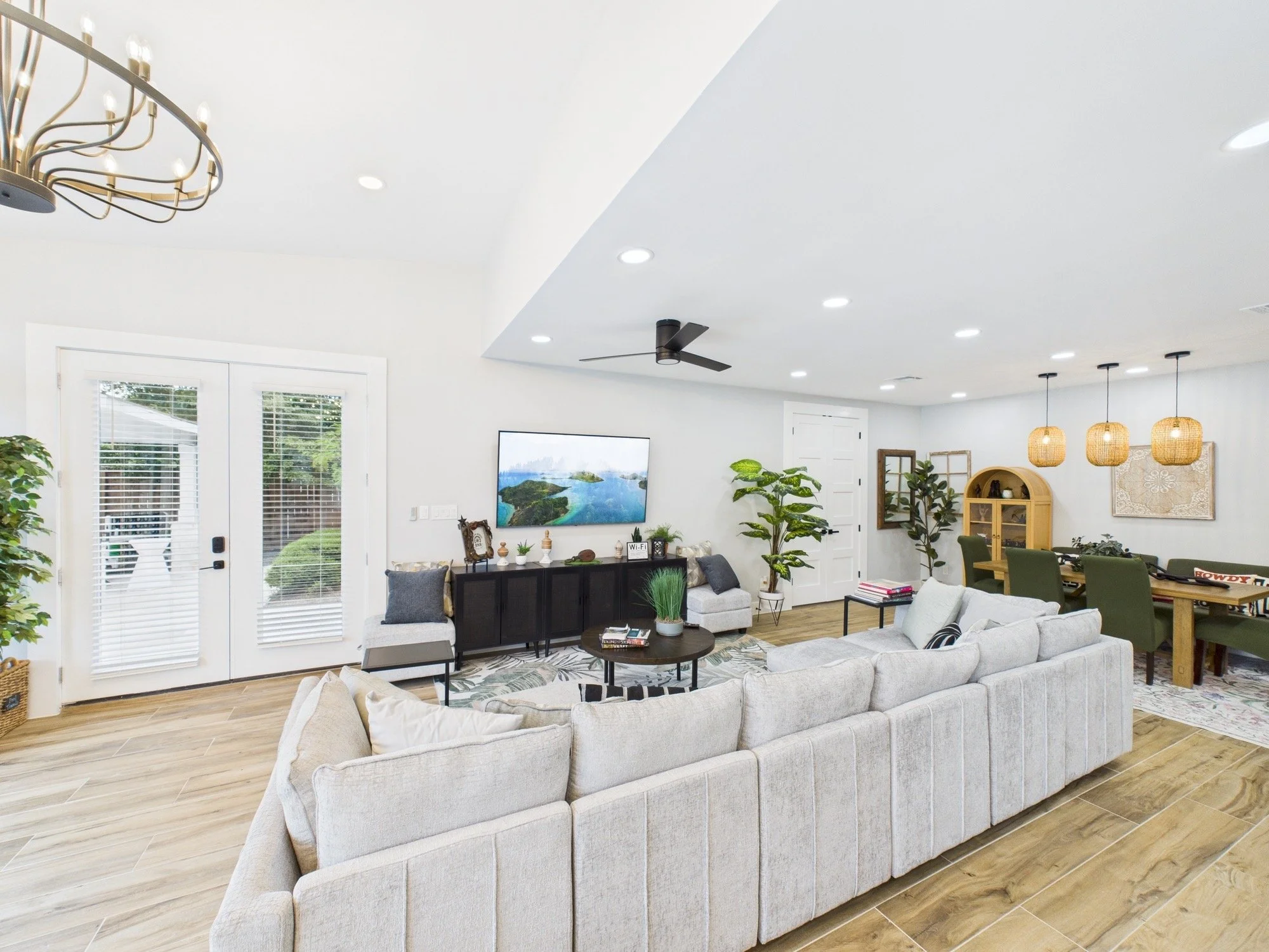 Bright living room with white walls, wooden flooring, a large L-shaped beige sofa, and a TV on the wall. Decor includes plants, framed artwork, a black sideboard, and a wooden dining table with green chairs. Rattan pendant lights and a ceiling fan ar