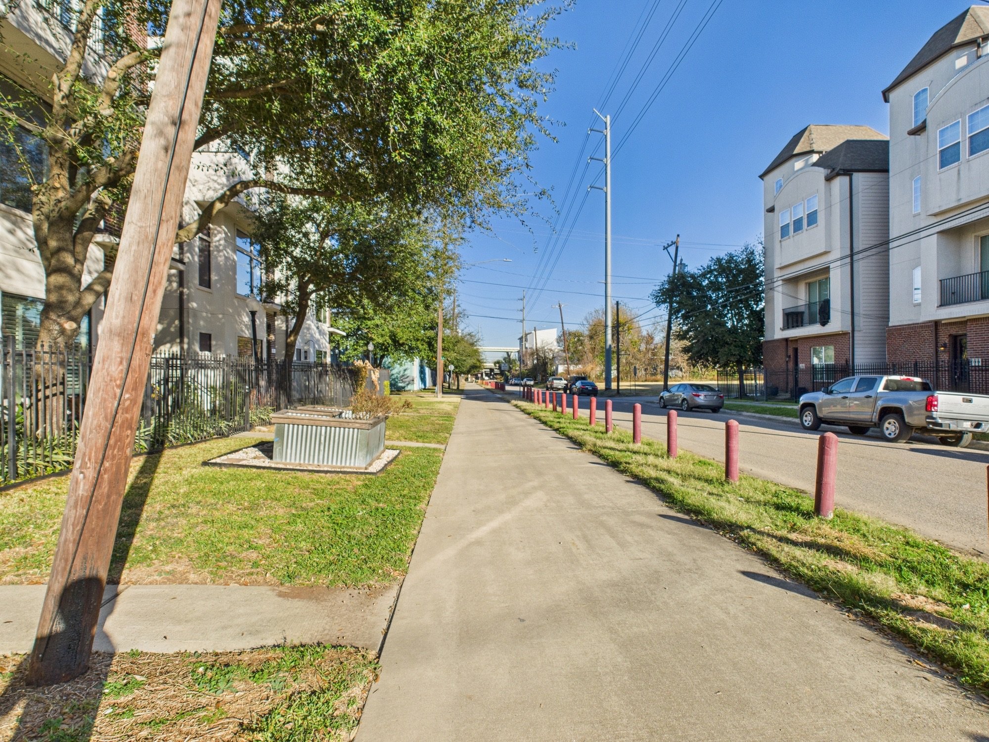 A sidewalk with pink posts on the right, a grassy area on the left, trees, multi-story apartment buildings, parked cars, utility poles, power lines, and a clear blue sky.