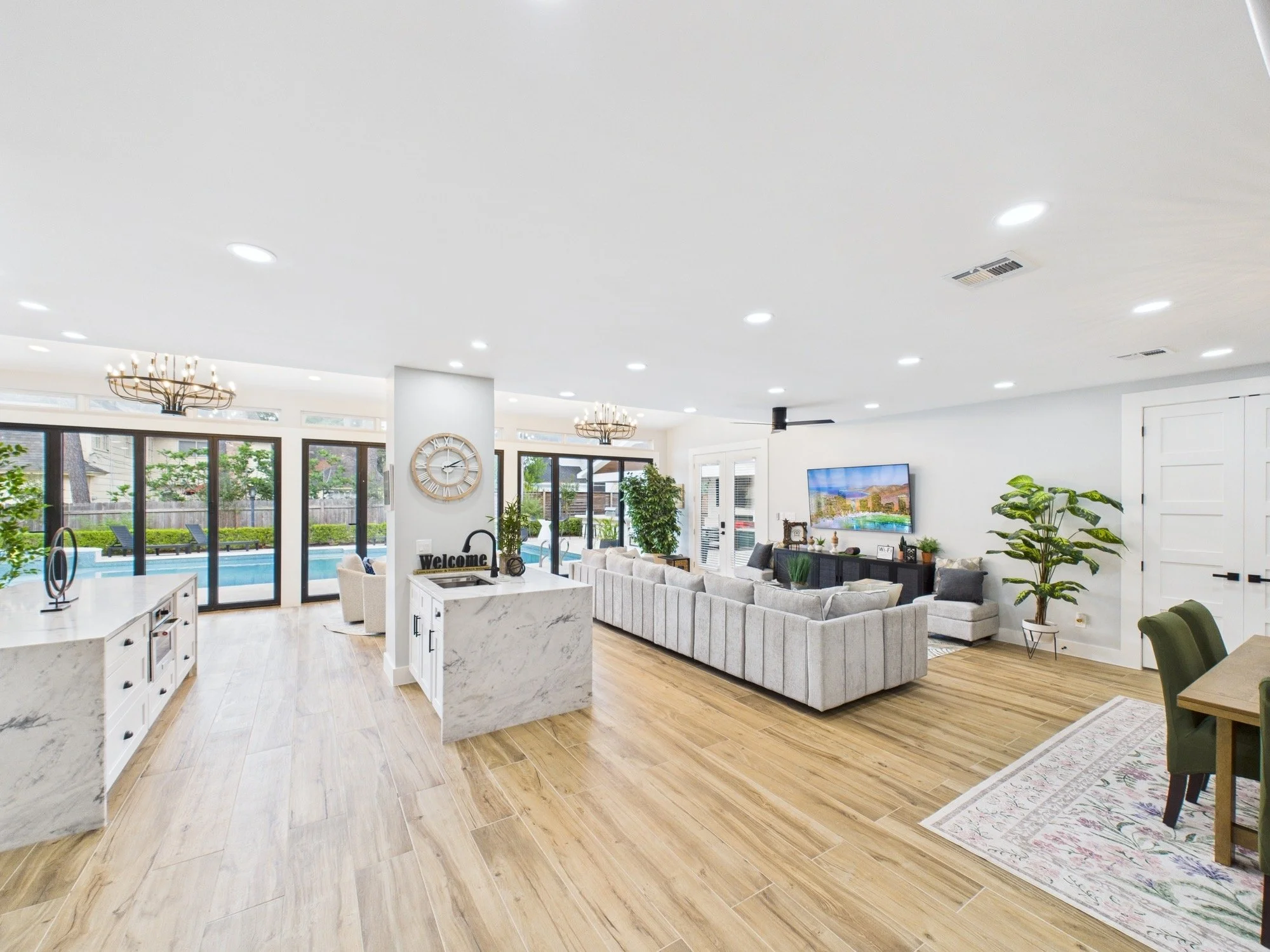 Bright living room with white ceiling, multiple recessed lights, large windows, gray sofa, wall clock, TV, potted plants, and wooden floor.