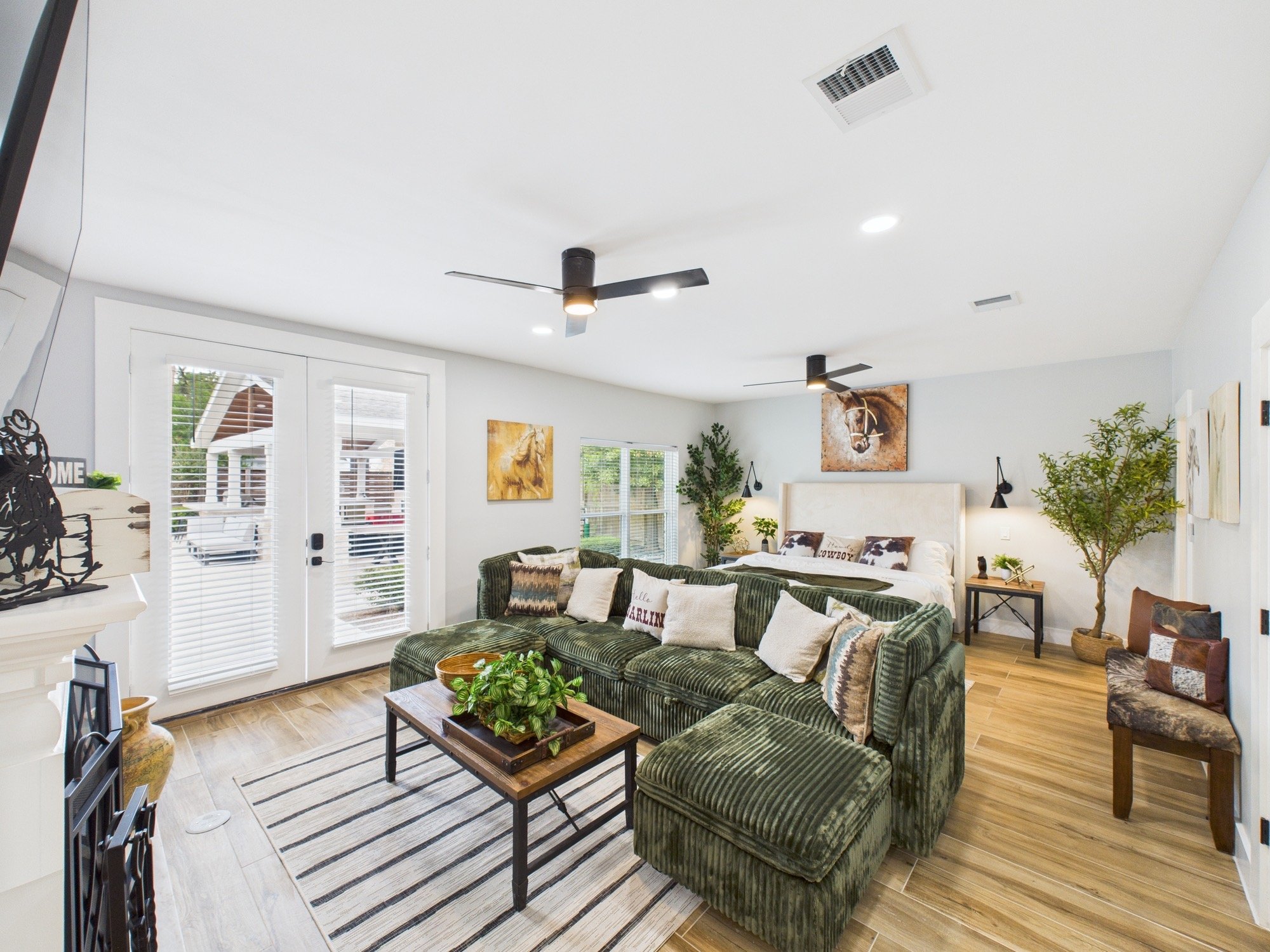 Living room with green velvet sofa, artwork of a horse and a cowgirl, wooden coffee table with a plant, and large windows with white blinds.