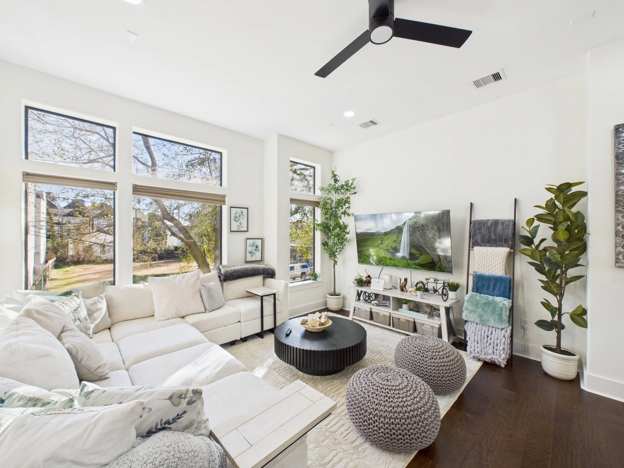 Bright living room with large windows, white sofas, a black round coffee table, green plants, framed wall art, a wall-mounted TV displaying a landscape, and various knitted poufs.