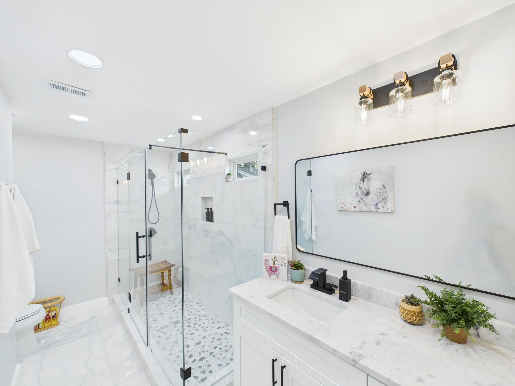 Modern bathroom with white and gray marble surfaces, glass shower enclosure, potted plants, and a large mirror.