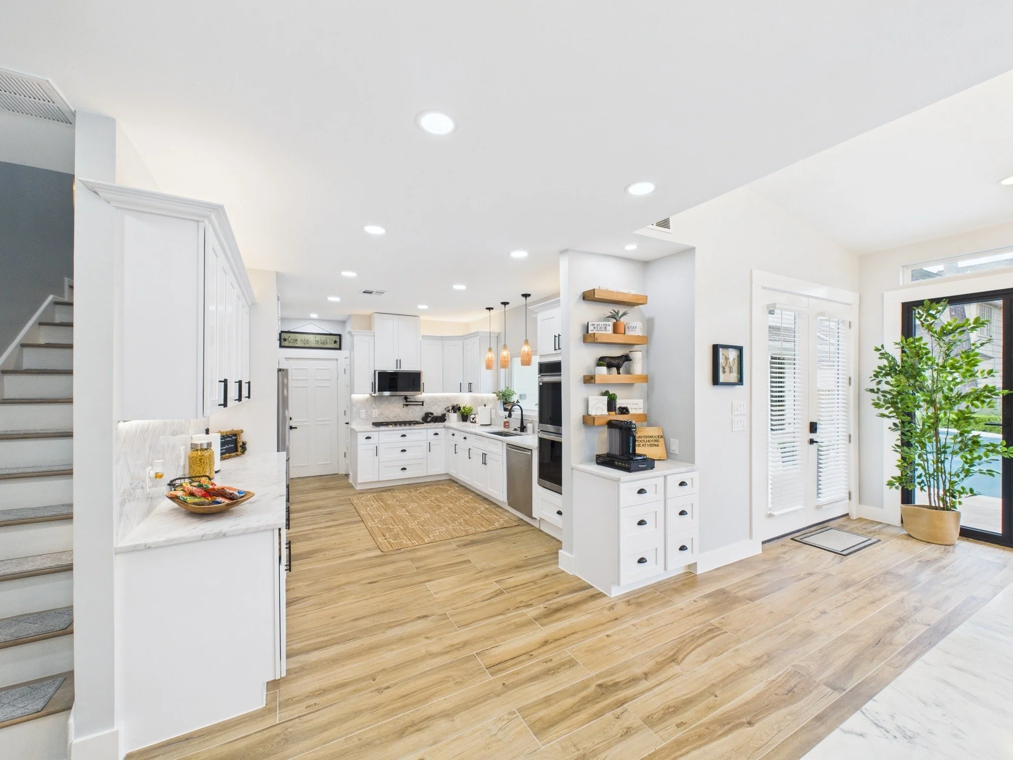 Bright, modern kitchen with white cabinets, wooden floors, a small island with a bowl of snacks, and a wall with open wooden shelves holding decorative items and potted plants. Large windows with blinds and a potted tree are visible.