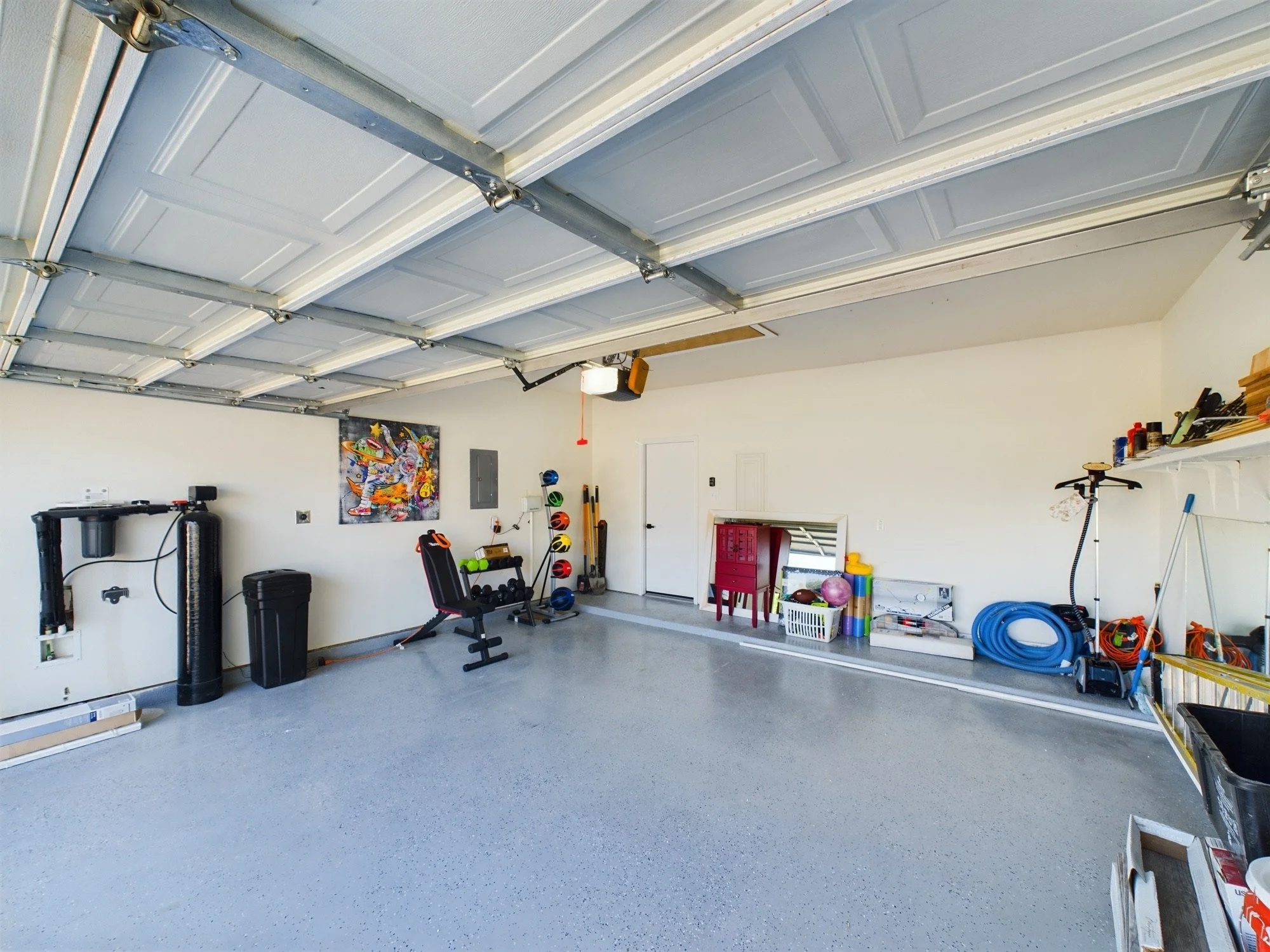 A spacious garage with an open ceiling and a closed metal garage door. The garage contains various workout equipment including a punching bag, a black exercise bench, a set of dumbbells, helmets, and a collection of colorful balls and miscellaneous i